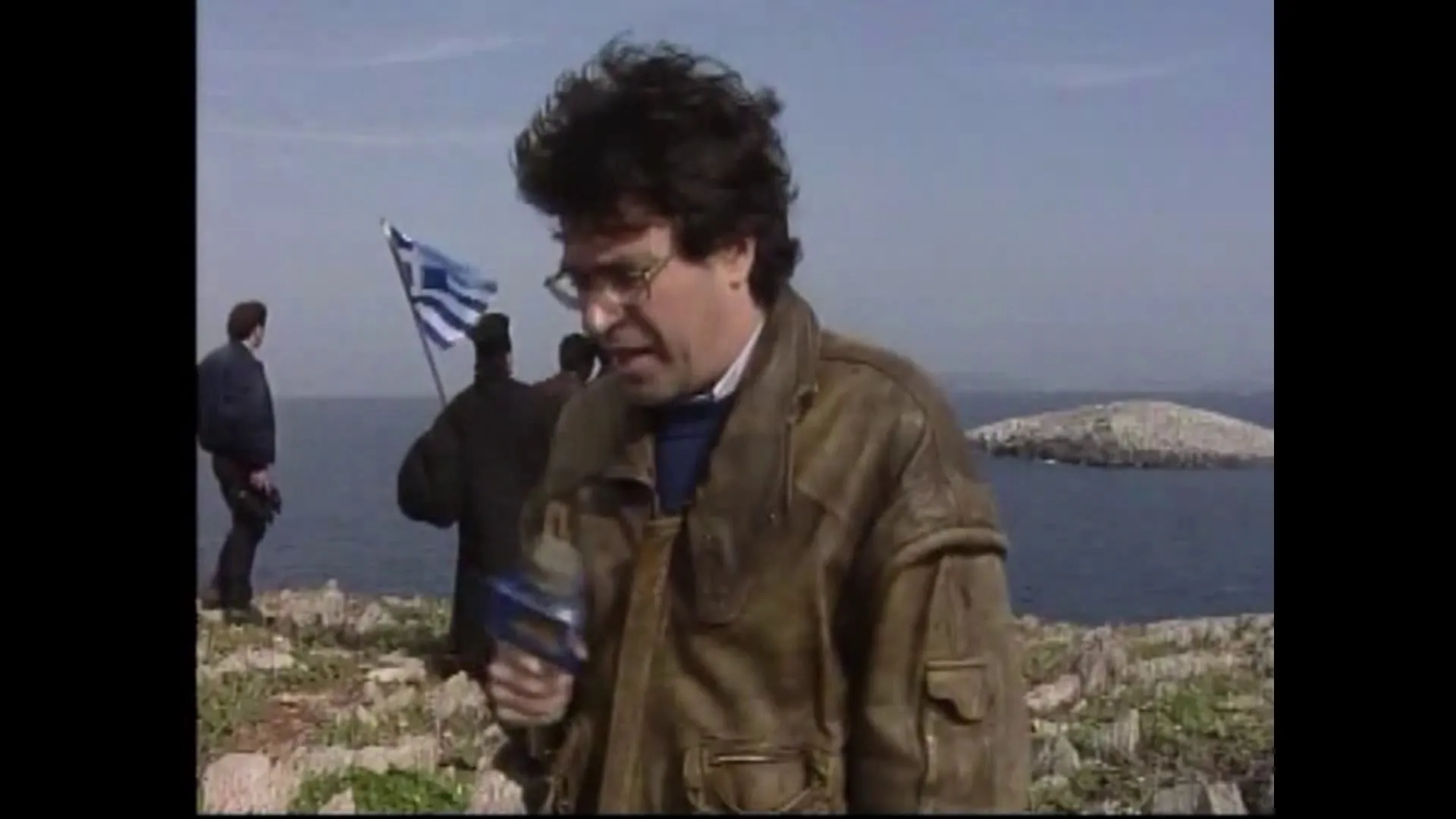 Ο Αργύρης Ντινόπουλος μιλά στη βραχώδη νησίδα κρατώντας μικρόφωνο του ΑΝΤ1, με δύο ακόμη άνδρες στο πλάνο· πίσω του η σημαία και δεύτερη βραχονησίδα στη θάλασσα.
