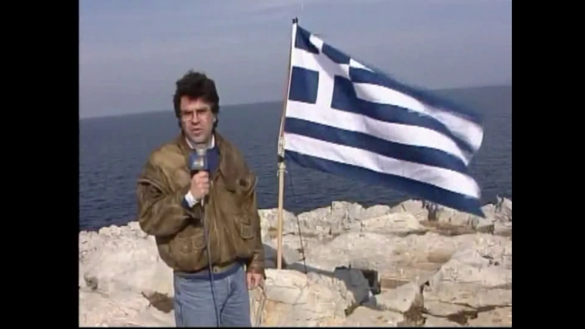 Ο Αργύρης Ντινόπουλος με μικρόφωνο του ΑΝΤ1 στα βράχια, δίπλα σε μεγάλη ελληνική σημαία που κυματίζει σε ιστό.