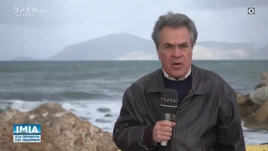Καρέ από τηλεοπτική μετάδοση του OPEN: ο Αργύρης Ντινόπουλος με μικρόφωνο σε βραχώδη ακτή με κύματα· κάτω αριστερά ο τίτλος «ΙΜΙΑ — ΣΤΑ ΠΡΟΘΥΡΑ ΤΟΥ ΠΟΛΕΜΟΥ».
