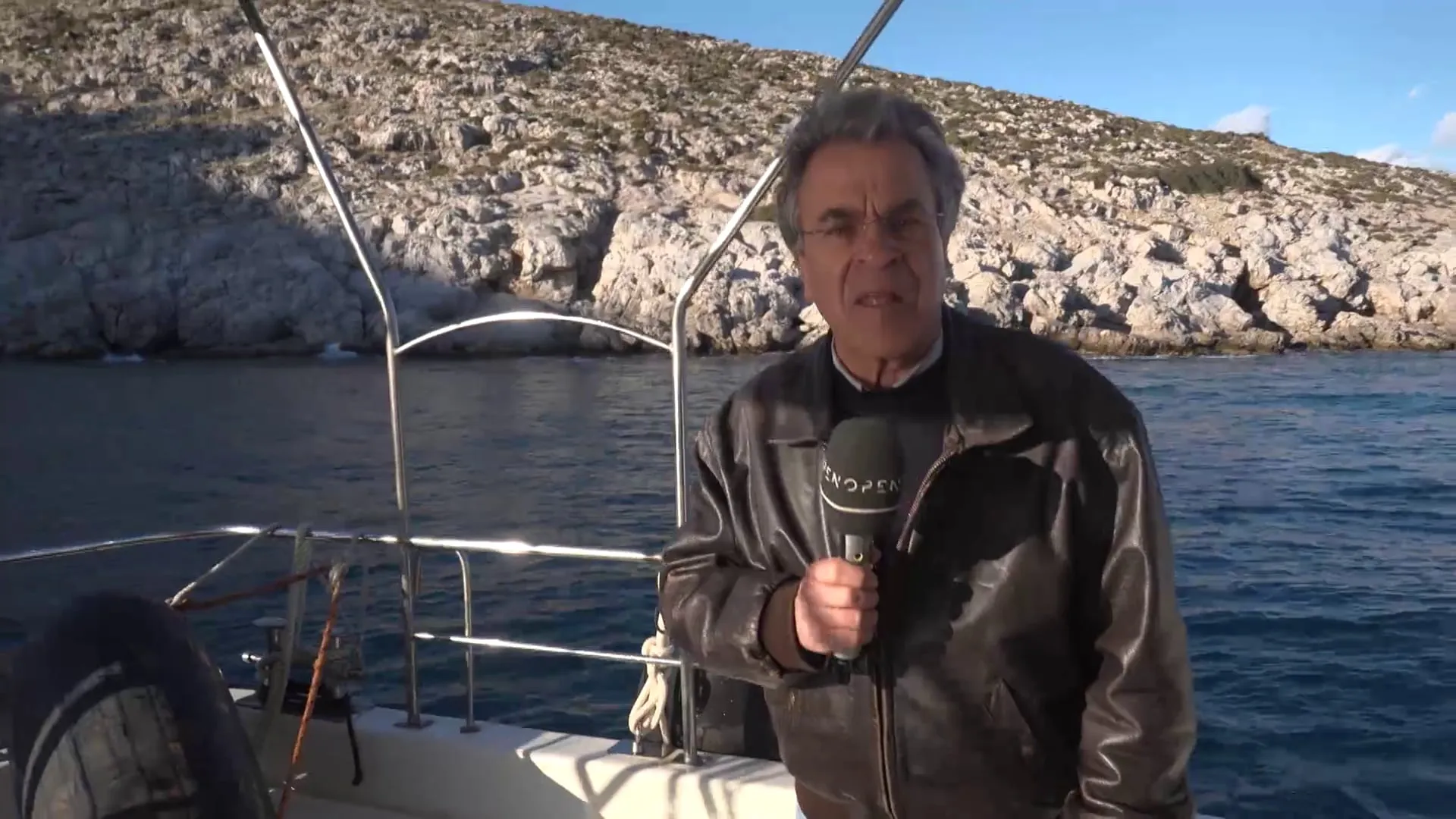 Ο Αργύρης Ντινόπουλος με μικρόφωνο σε σκάφος με ανοξείδωτα κάγκελα· πίσω του η βραχώδης ακτή της νησίδας.