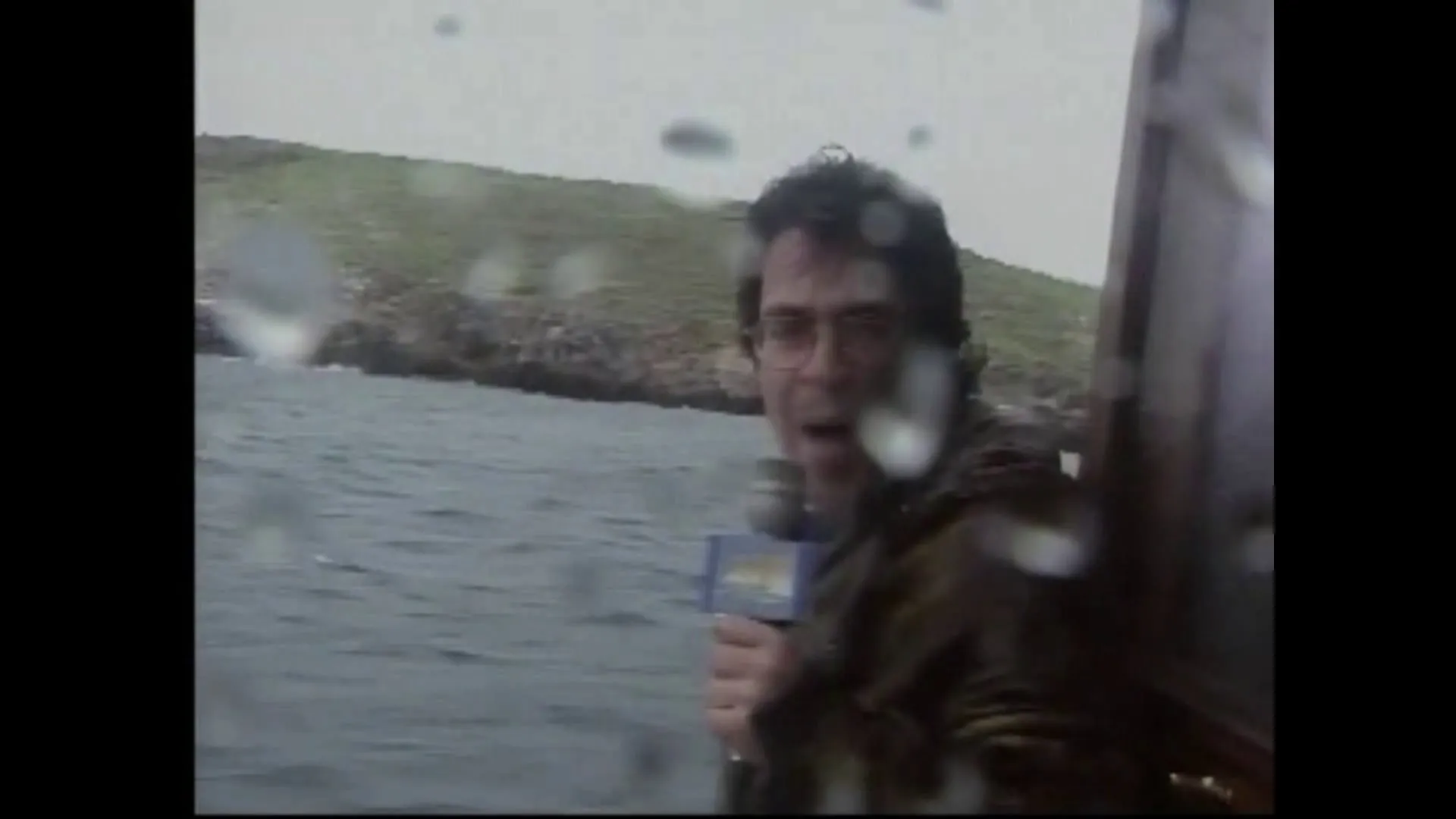 Ο Αργύρης Ντινόπουλος σε σκάφος με σταγόνες θαλασσινού νερού στον φακό, με τη βραχονησίδα πίσω του.