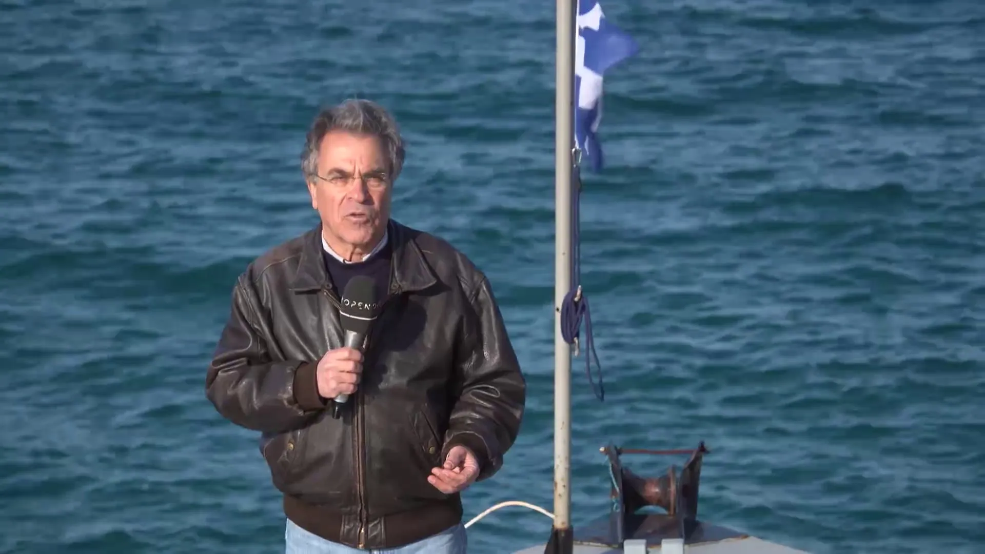 Ο Αργύρης Ντινόπουλος με μικρόφωνο σε σκάφος, με ελληνική σημαία σε κοντάρι δίπλα του και ανοιχτή θάλασσα πίσω.