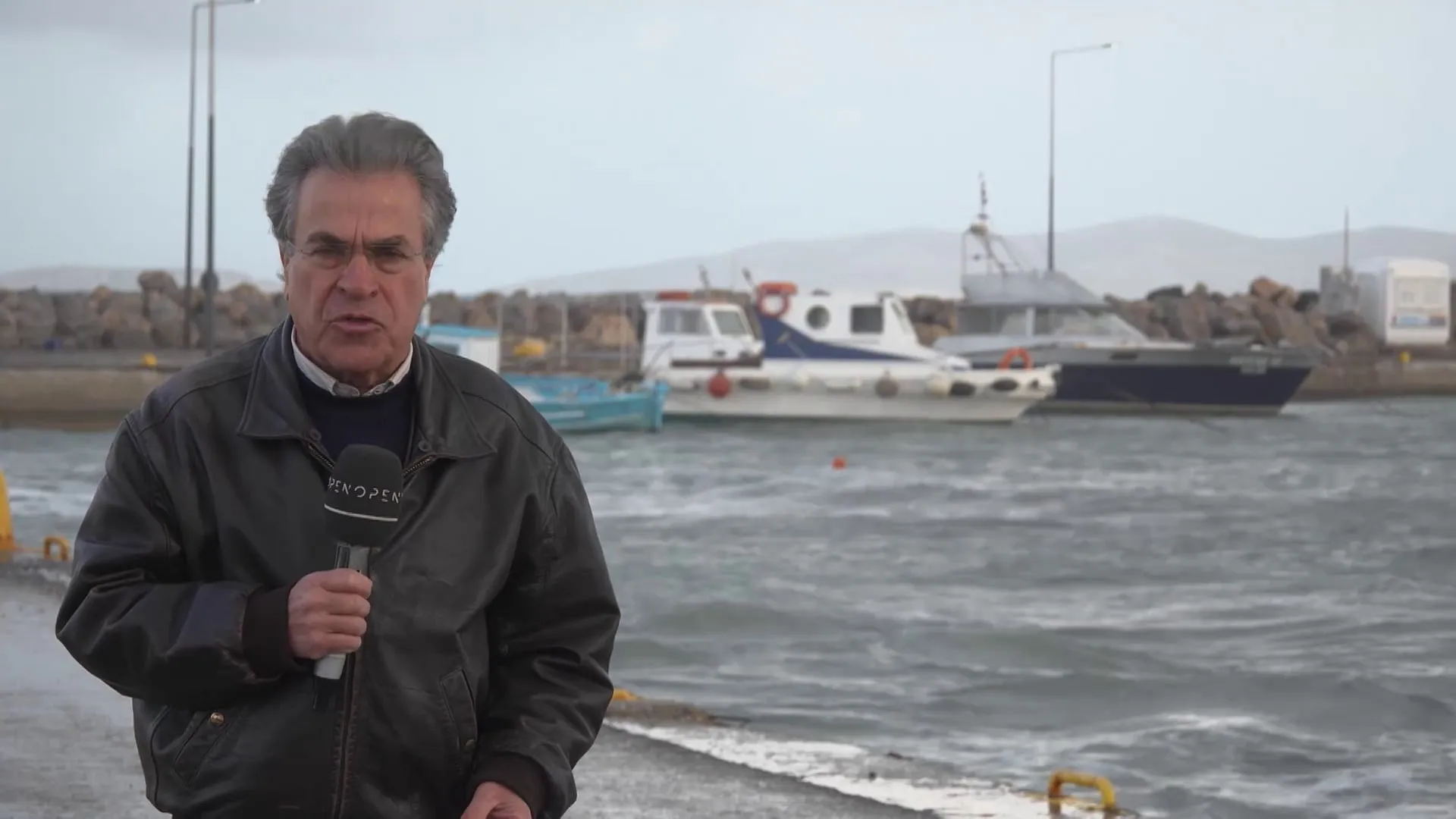Ο Αργύρης Ντινόπουλος με μικρόφωνο σε προκυμαία της Καλύμνου, με δεμένα σκάφη και φουρτουνιασμένη θάλασσα πίσω του.