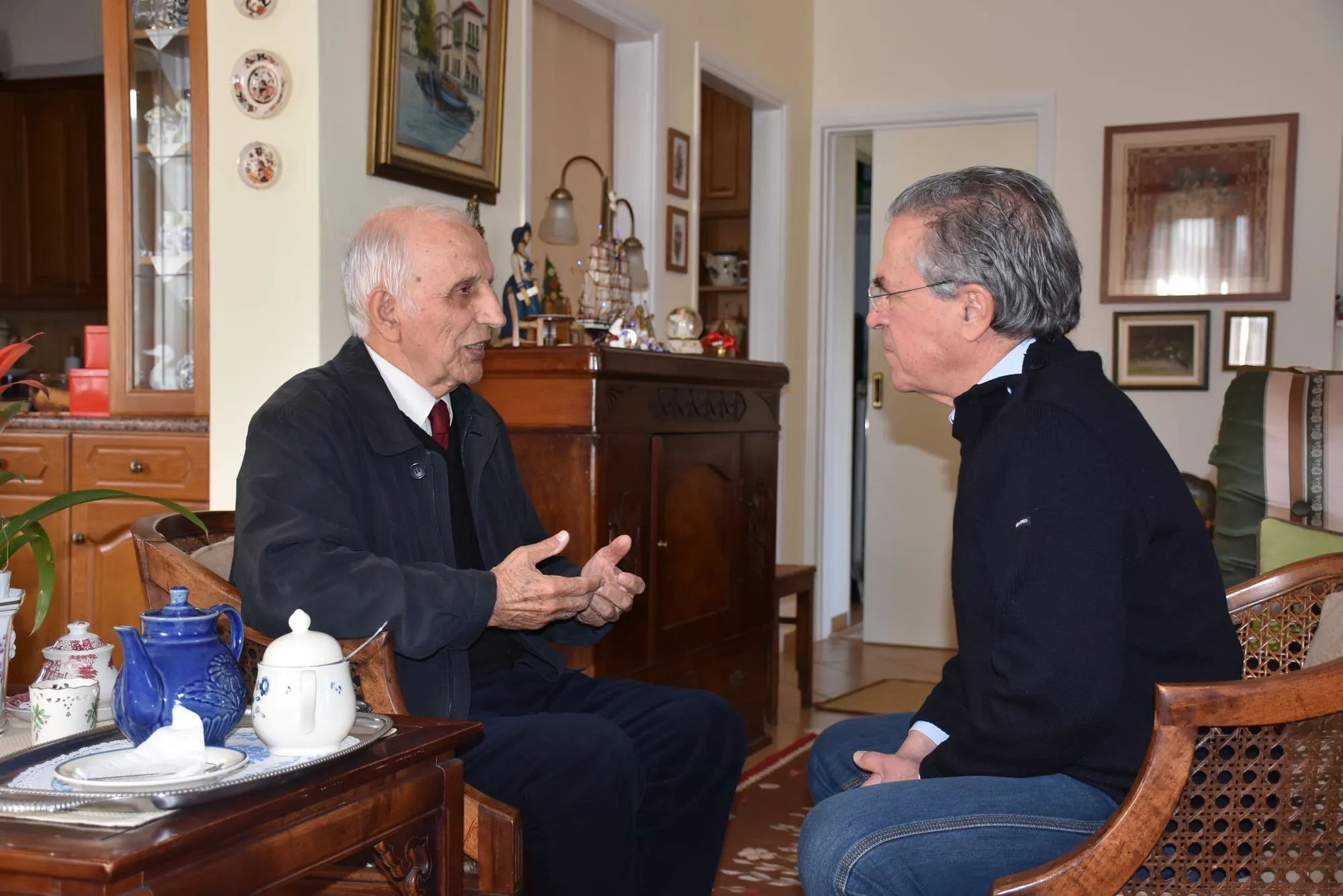 Ηλικιωμένος άνδρας με σκούρο μπουφάν, λευκό πουκάμισο και σκούρα κόκκινη γραβάτα μιλά καθισμένος σε σαλόνι σπιτιού, με τον Αργύρη Ντινόπουλο καθισμένο απέναντί του.