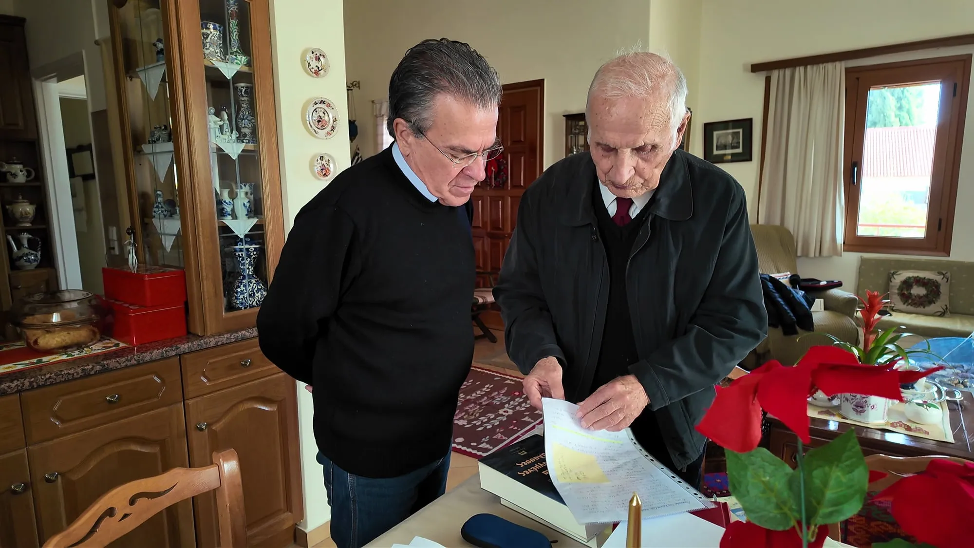 Δύο άνδρες όρθιοι πάνω από τραπέζι, ο ένας κρατά και δείχνει ένα έγγραφο· δίπλα ένα βιβλίο και ένα κόκκινο αλεξανδρινό.