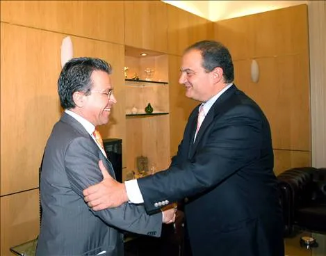 Δύο άνδρες με σκούρα κοστούμια ανταλλάσσουν χειραψία σε χώρο με ξύλινη επένδυση· ο ένας κρατά τον άλλον από τον ώμο και πίσω τους διακρίνονται ράφια με μικρά διακοσμητικά.