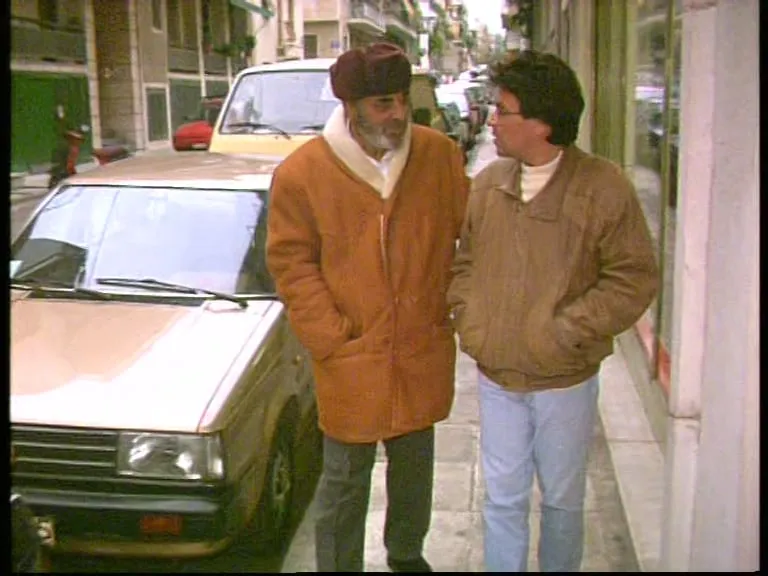 Two men walk along a narrow city pavement in winter clothes. At left Stelios Kazantzidis, in a dark red knitted cap, bearded, in a brown coat with a white-lined collar; at right Argyris Dinopoulos, in glasses and a pale jacket, turned towards him. Parked cars and shopfronts beside them; the street recedes behind.