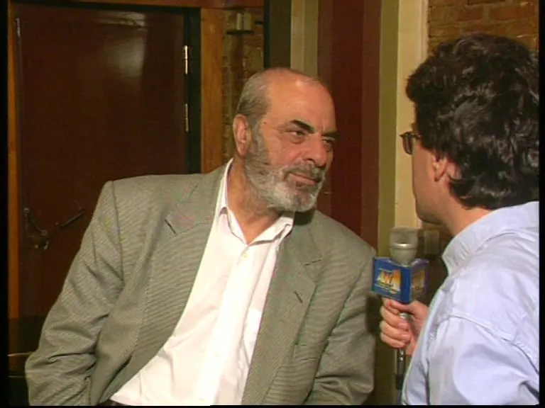 An interior with a brick wall and dark wooden doors. Stelios Kazantzidis is seated in a grey jacket over an open white shirt, grey-bearded, looking to the right. At the right edge of the frame, his back to the camera, Argyris Dinopoulos in a pale blue shirt holds a microphone with an Antenna flag.