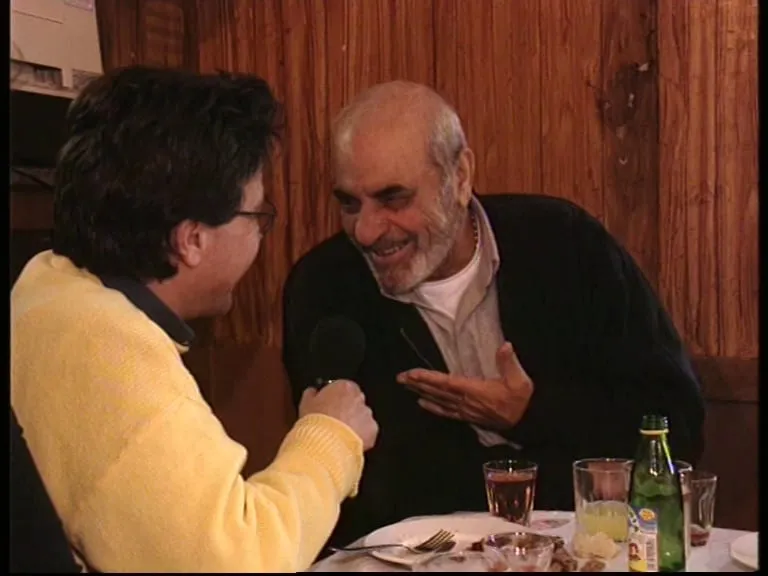 A taverna interior with wood-panelled walls. At right Stelios Kazantzidis, in a dark cardigan over a white polo-neck, smiles broadly with one hand raised, palm open. At left, three-quarters from behind, Argyris Dinopoulos in a yellow jumper holds a microphone. On the table in front of them, glasses, a green bottle and cutlery.