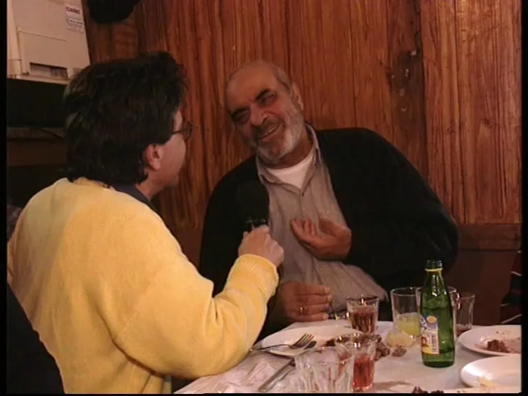 The same taverna table. Stelios Kazantzidis smiles with one hand at his chest; opposite him Argyris Dinopoulos, in a yellow jumper, holds the microphone close to him. On the counter behind at left, a white appliance; on the table, glasses, a green bottle and plates.
