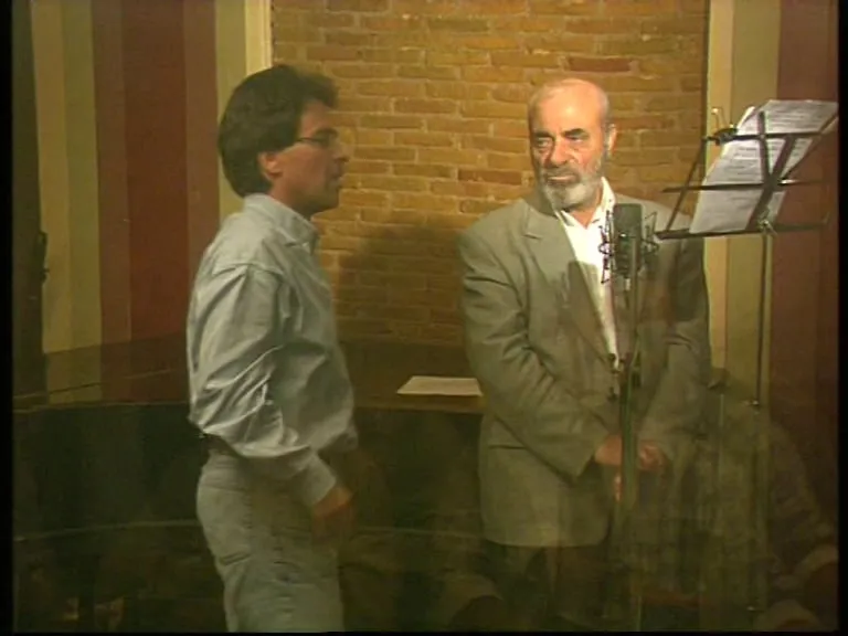 A recording room with a brick wall, a stand holding sheet music and a microphone on a stand. At right Stelios Kazantzidis stands in a grey jacket and white shirt; at left, his back to the camera, Argyris Dinopoulos in a pale blue shirt. At the left edge of the frame the black lid of a piano is visible.