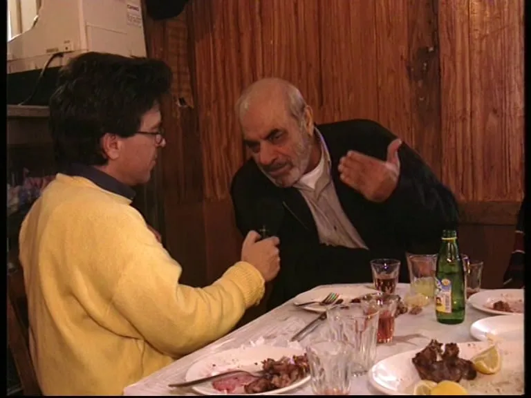 A wider view of the same table. Stelios Kazantzidis speaks with one hand raised; opposite him Argyris Dinopoulos in a yellow jumper with the microphone. On the table, plates with the remains of a meal, glasses, a green bottle and a wedge of lemon.