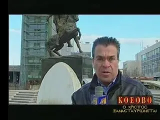 Ο Αργύρης Ντινόπουλος μιλά στην κάμερα κρατώντας μπλε μικρόφωνο με χρυσαφί σήμα, με χάλκινο έφιππο ανδριάντα σε ψηλό βάθρο πίσω του και σύγχρονα κτίρια της πόλης γύρω. Κάτω δεξιά, ο ένθετος τίτλος της εκπομπής του «ΚΟΣΟΒΟ / Ο ΧΡΙΣΤΟΣ ΞΑΝΑΣΤΑΥΡΩΝΕΤΑΙ».