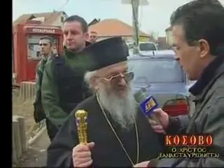Ορθόδοξος επίσκοπος με μαύρο ράσο και μαύρο κυλινδρικό καλυμμαύχι, λευκή γενειάδα και γυαλιά, κρατά χρυσή ποιμαντική ράβδο και μιλά σε μπλε μικρόφωνο με χρυσαφί σήμα που του κρατά άνδρας γυρισμένος πλάτη στον φακό· πίσω τους άνδρες, κόκκινο περίπτερο και σπίτια με κεραμοσκεπές. Κάτω δεξιά, ο ένθετος τίτλος της εκπομπής του «ΚΟΣΟΒΟ / Ο ΧΡΙΣΤΟΣ ΞΑΝΑΣΤΑΥΡΩΝΕΤΑΙ».
