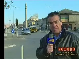 Ο Αργύρης Ντινόπουλος μιλά στην κάμερα με μικρόφωνο του ΑΝΤ1 σε δρόμο πόλης· πίσω του μιναρές και ο τρούλος τζαμιού, χαμηλά κτίρια, φανάρια, ένα κίτρινο φορτηγάκι και αυτοκίνητα σε διασταύρωση. Κάτω δεξιά, ο ένθετος τίτλος της εκπομπής του «ΚΟΣΟΒΟ / Ο ΧΡΙΣΤΟΣ ΞΑΝΑΣΤΑΥΡΩΝΕΤΑΙ».
