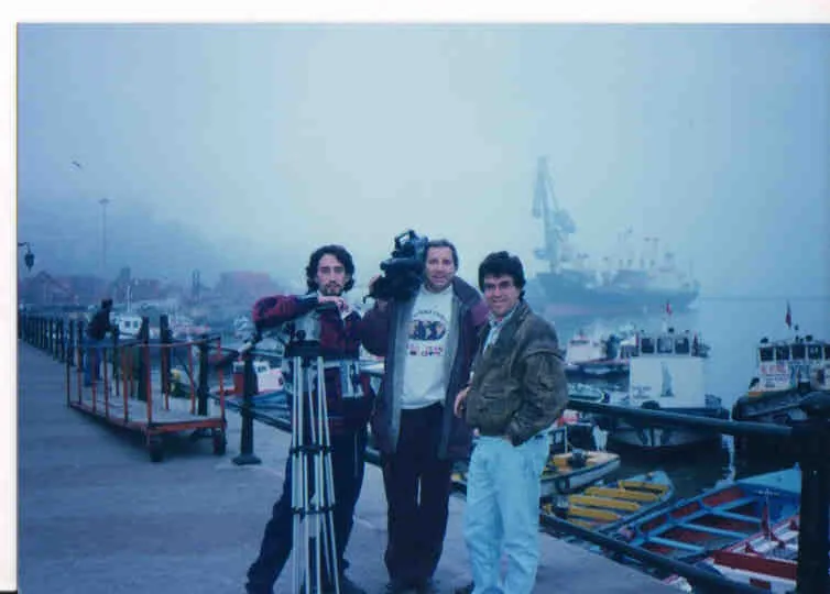 Three men standing at the edge of a quay in thick mist; the one on the left, in a maroon jacket, carries a television camera on his shoulder with an opened tripod in front of him, the one in the middle in a white printed sweatshirt steadies the camera, and the one on the right, in a beige jacket and pale jeans, looks at the lens; behind them dozens of small open boats moored in rows, a metal railing running the length of the quay, a harbour crane and a cargo ship in the mist.