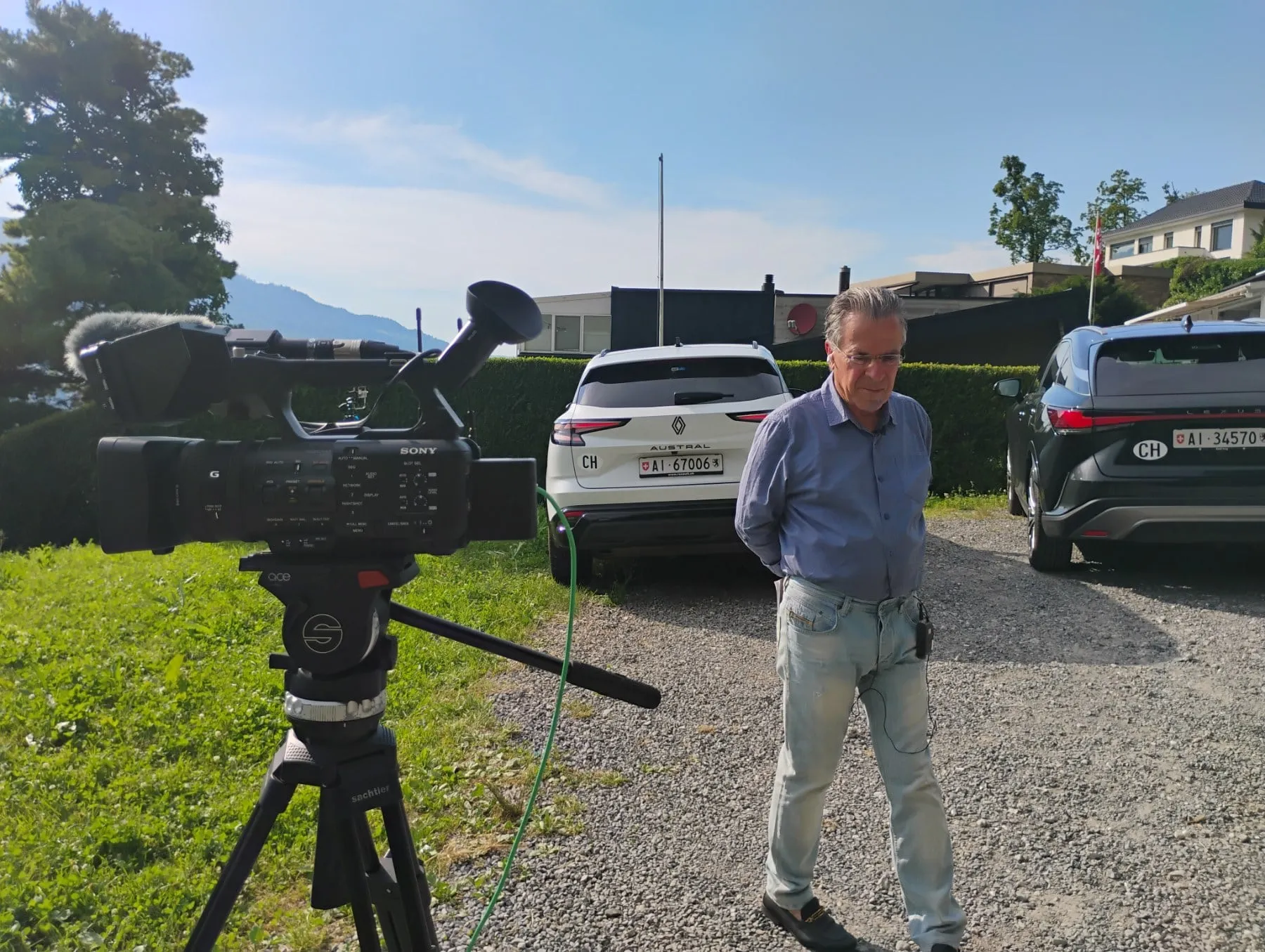 A television camera on a tripod at the left of the frame, its lens pointing out of the picture; to its right Argyris Dinopoulos walks across a gravel forecourt, looking down, in a blue shirt and pale jeans, a small black device clipped at his belt with a cable hanging from it; behind him two parked cars with Swiss number plates, a clipped hedge, low buildings, a red flag on a pole to the right and a mountain beyond.
