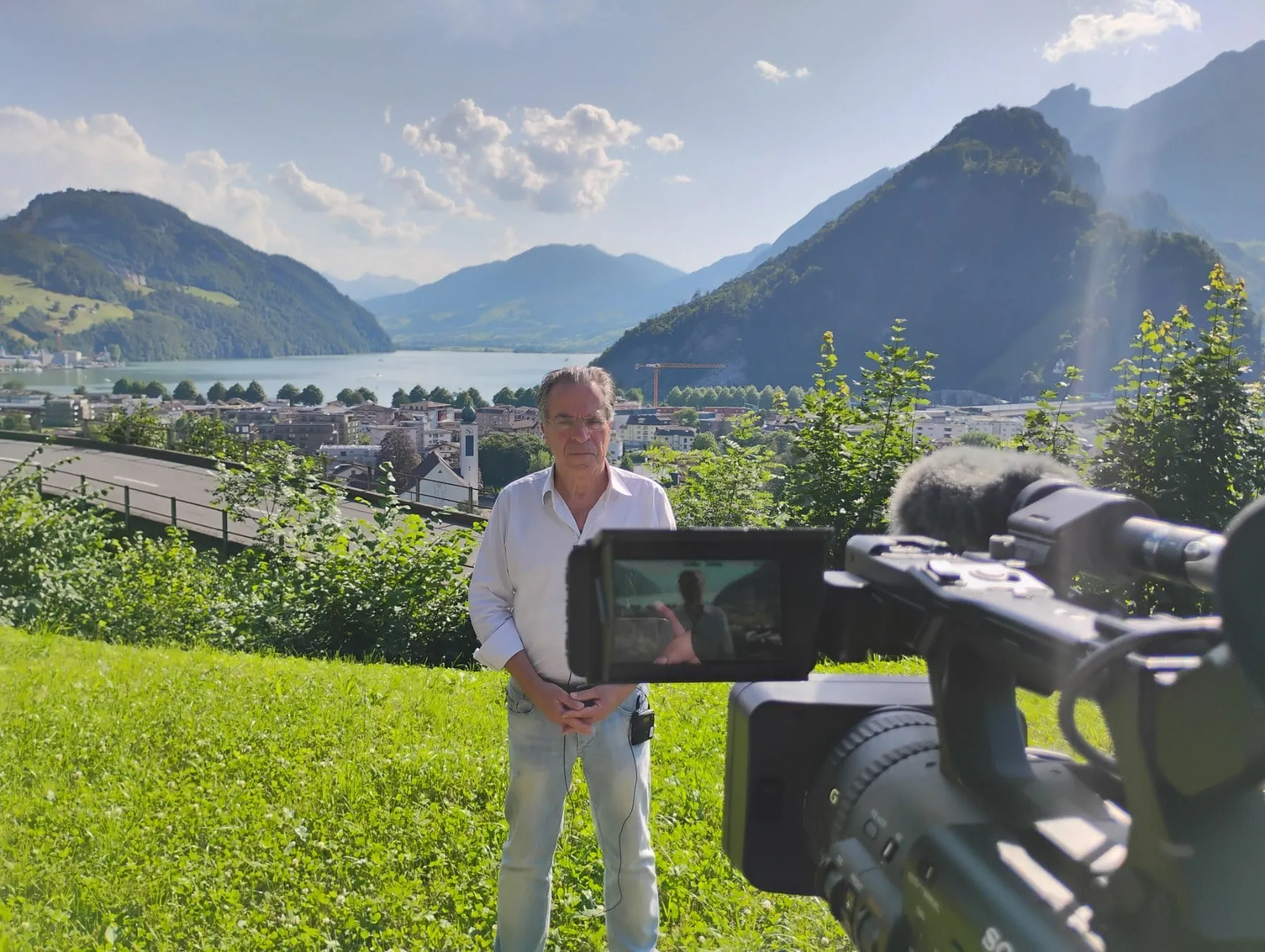 Argyris Dinopoulos standing on a grassy slope in a white shirt and pale jeans, hands clasped in front of him; behind him a lake between mountains, the town on the shore and a road curving away to the left; in the right foreground a television camera with its monitor swung out.