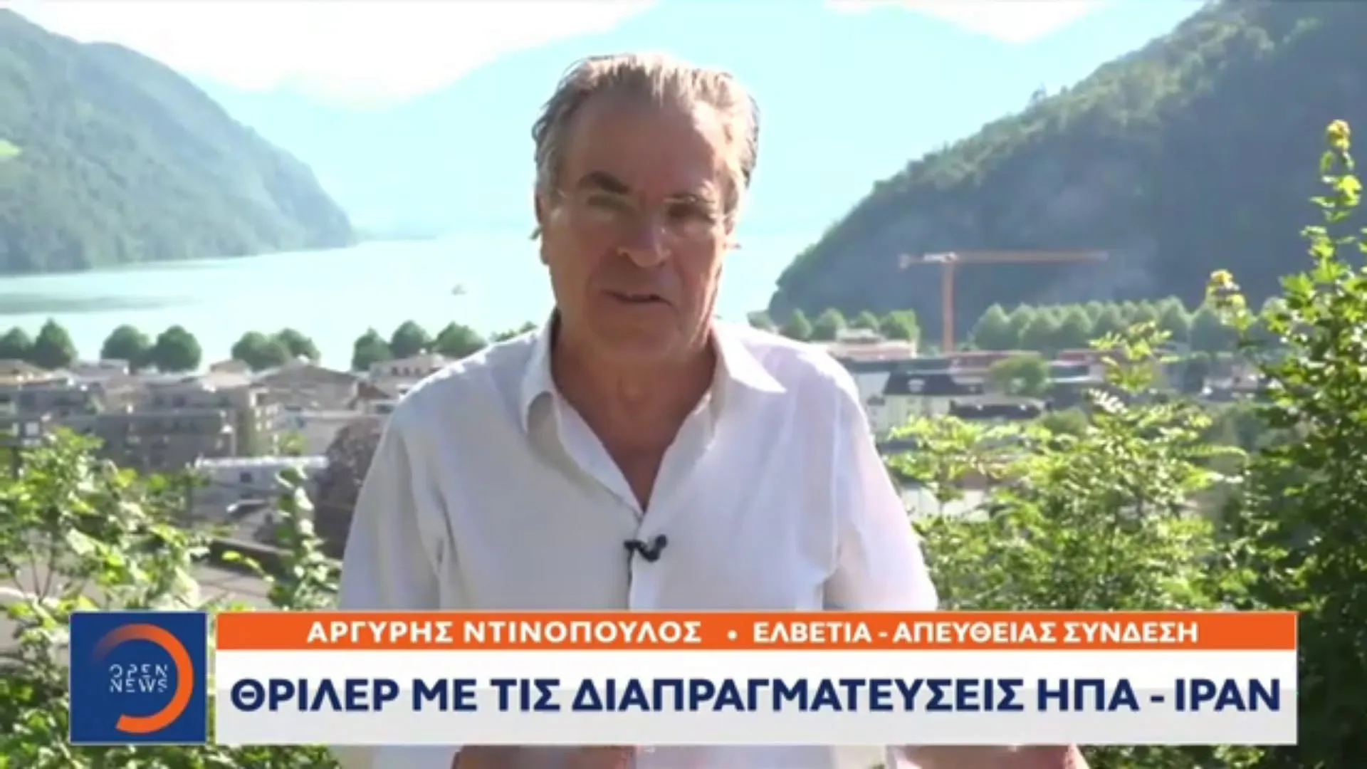 Frame from an OPEN television broadcast — Argyris Dinopoulos speaking to camera in a white shirt with a clip-on microphone, the lake, the mountains and the town behind him; lower left the OPEN NEWS logo, beside it his name and «ΕΛΒΕΤΙΑ – ΑΠΕΥΘΕΙΑΣ ΣΥΝΔΕΣΗ» — «Switzerland — live» — and below it the headline «ΘΡΙΛΕΡ ΜΕ ΤΙΣ ΔΙΑΠΡΑΓΜΑΤΕΥΣΕΙΣ ΗΠΑ – ΙΡΑΝ» — «Thriller in the US–Iran negotiations».