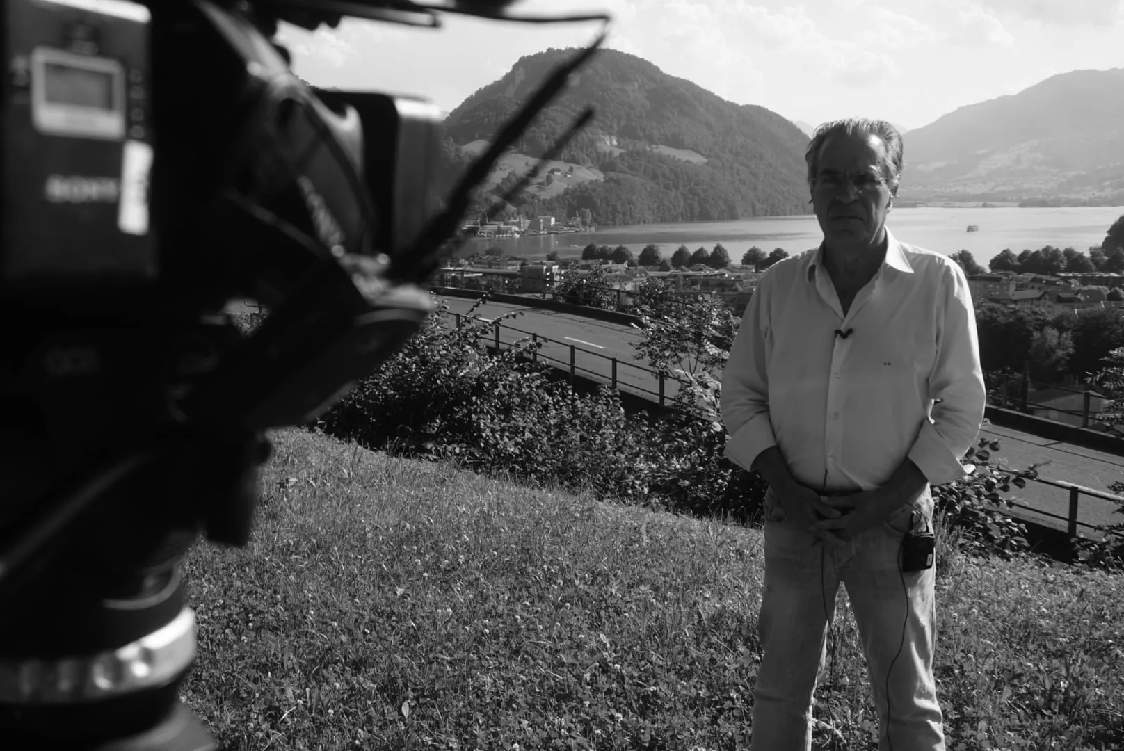 Black-and-white photograph — in the left foreground, blurred and very close to the lens, a television camera; to the right Argyris Dinopoulos in a white shirt, hands clasped in front of him, at the edge of a grassy slope; behind him shrubs, a road with a guard rail, the town on the shore and the lake between mountains.