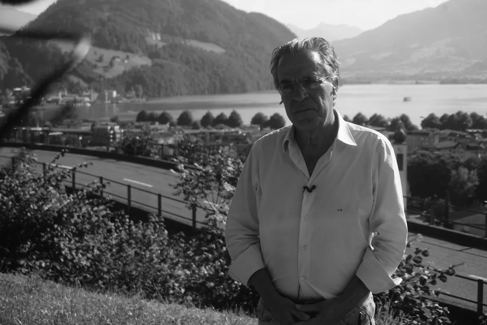 Ασπρόμαυρη φωτογραφία — ο Αργύρης Ντινόπουλος σε κοντινό πλάνο, με λευκό πουκάμισο, γυαλιά και μικρόφωνο πέτου, κοιτάζοντας ελαφρώς προς τα κάτω· πίσω του δέντρα, δρόμος με κιγκλίδωμα, η πόλη στην όχθη με έναν πύργο με ρολόι και βολβοειδή απόληξη, και η λίμνη ανάμεσα σε βουνά· στην πάνω αριστερή γωνία ένα θολό σκούρο σχήμα, πολύ κοντά στον φακό.