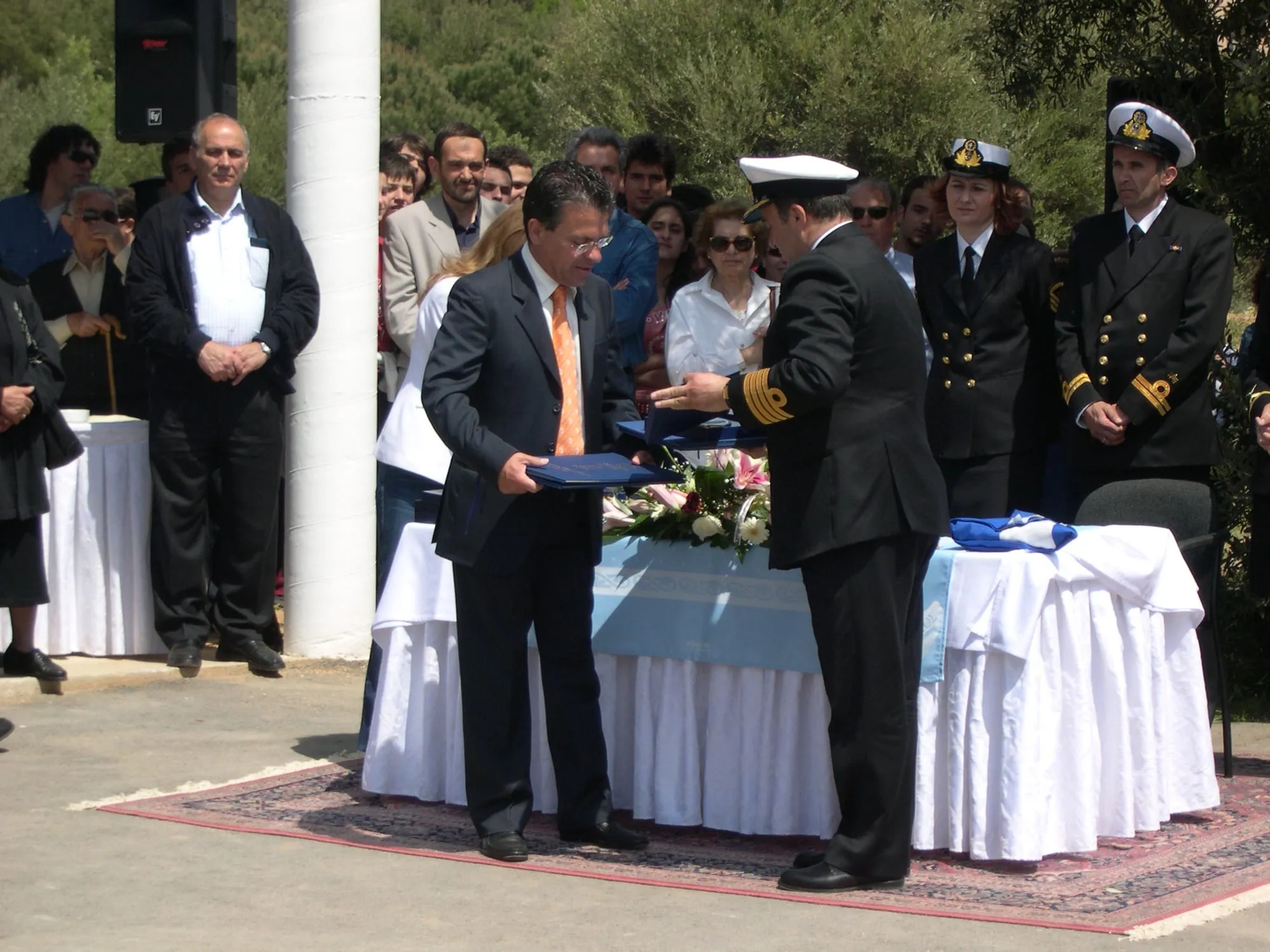 Υπαίθρια τελετή: ο Αργύρης Ντινόπουλος, με σκούρο κοστούμι και πορτοκαλί γραβάτα, κρατά μπλε φάκελο και τον ανταλλάσσει με αξιωματικό του Πολεμικού Ναυτικού με επίσημη στολή και λευκό πηλήκιο. Δεξιά στέκονται δύο ακόμη αξιωματικοί· πίσω, πλήθος κόσμου και ελιές. Στο τραπέζι με το γαλάζιο κάλυμμα, λουλούδια και διπλωμένη ελληνική σημαία.