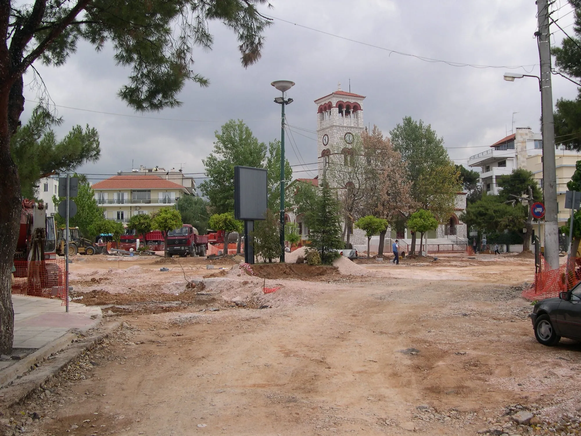 Η κεντρική πλατεία σε εργοτάξιο: γυμνό χώμα και μπάζα σε όλη την έκταση, εκσκαφέας και ανατρεπόμενα φορτηγά αριστερά, πορτοκαλί πλέγμα ασφαλείας, ένας εργάτης περπατά στο βάθος. Πίσω, η πέτρινη εκκλησία με το ψηλό καμπαναριό και το ρολόι, πολυκατοικίες και συννεφιασμένος ουρανός.