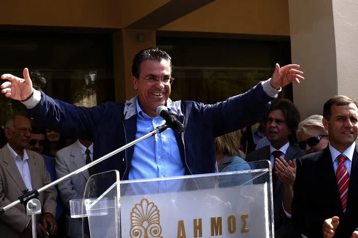 Ο Αργύρης Ντινόπουλος σε βήμα με την επιγραφή «ΔΗΜΟΣ», με υψωμένα χέρια προς πλήθος που επευφημεί.