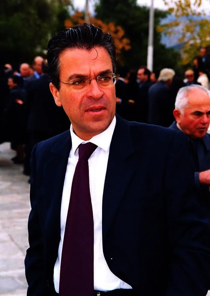 Κοντινό πορτρέτο του Αργύρη Ντινόπουλου με κοστούμι και γυαλιά, ανάμεσα σε πλήθος επίσημης εκδήλωσης.