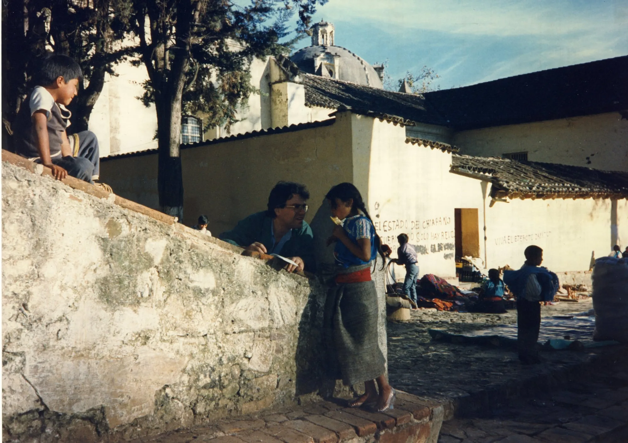 At the left, a low stone wall with a boy sitting on top of it. Argyris Dinopoulos, in glasses and a green shirt over a blue one, rests his arms on the wall holding folded white papers. Facing him, standing on steps, a girl in a blue embroidered blouse, a wide red sash, a long grey skirt of heavy weave and silver sandals, raising something pale yellow to her mouth. Behind them whitewashed buildings with pantiled roofs and a dome with a lantern; painted in black on the wall, «EL ESTADO DE CHIAPA NO» and «SOLO HAY REVO», and further right, faded, «VIVA EL EJERCITO ZAPAT…», its last word trailing away. In the middle distance, at the foot of the wall, a child in a pale top and jeans, bent forward. At the right, a cobbled square with cloth goods spread on the ground, people sitting among them, sacks, and a boy with a blue bundle over his shoulder.