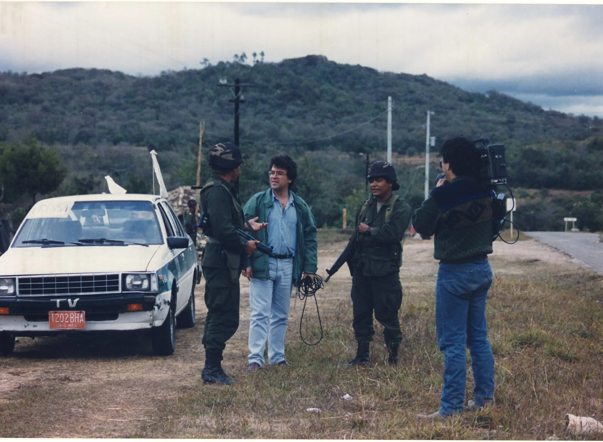 Argyris Dinopoulos stands on a grassy verge beside a metalled road, in a green jacket over a blue shirt, a belt and pale jeans; his right hand is open in a gesture, his left holds a coil of black cable. On either side of him two men in olive uniforms, camouflage-covered helmets and boots, each with a rifle; a third figure in the same uniform can be made out further back. At the left, a white car with a blue-green stripe: the word «PRENSA» across the top of the windscreen, «TV» on the bumper, a red number plate reading «1202 BHA» with «CHIS MEX» beneath it, and two white cloths tied high on the roof. At the right, his back to the lens, a man in a knitted jumper and jeans carries a television camera on his shoulder, a cable hanging from it. Behind, a scrub-covered hill, poles with wires and a clouded sky.