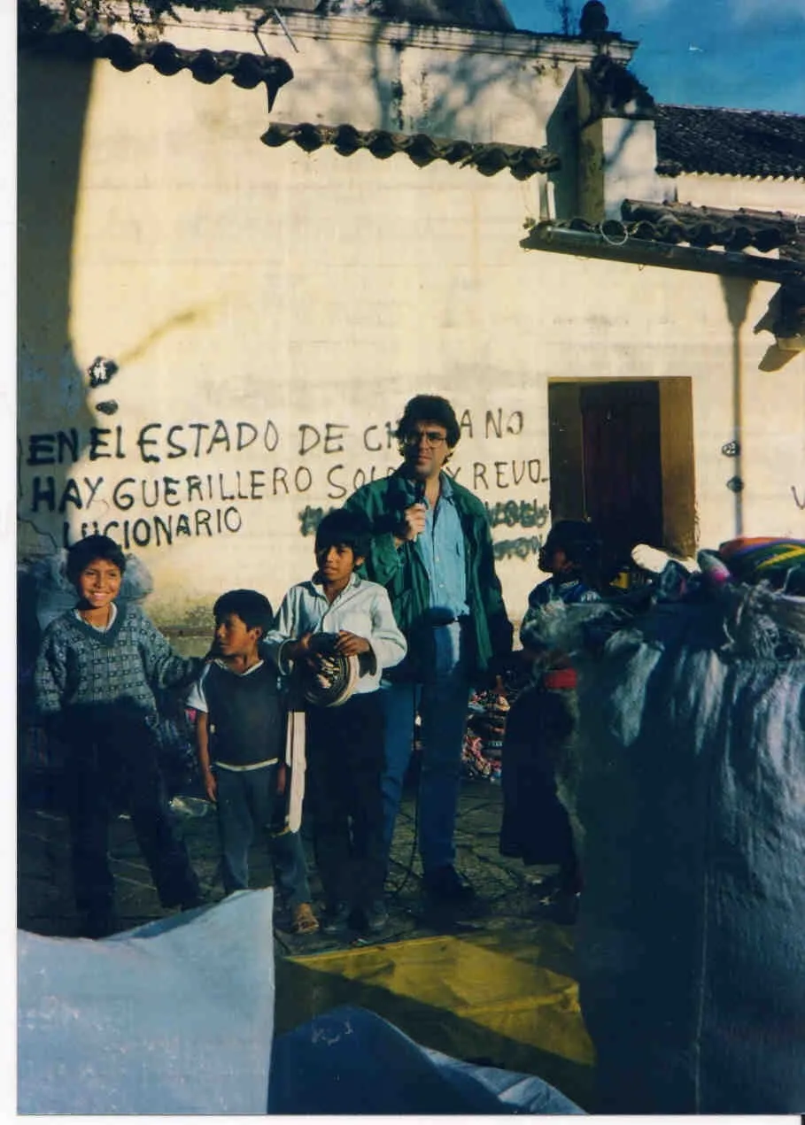 Upright frame. Argyris Dinopoulos stands in front of a whitewashed wall holding a microphone close to his face, in a green jacket over a blue shirt and jeans; a cable runs from the microphone down to the ground. At the left of the frame three children — one in a knitted jumper, one in a dark sleeveless top, and one in a white shirt holding a dark, spirally wound object with a pale woven strip hanging from it. On the wall behind them, painted in black capitals across three lines, «EN EL ESTADO DE CHIAPA NO HAY GUERILLERO SOLO HAY REVOLUCIONARIO» — «in the state of Chiapa there is no guerrilla, only revolutionary» — his head covering part of the first line and his shoulder part of the second, and lower down to their right an illegible slogan in green paint. At the right, a wooden door, pantiled roofs and a figure in dark clothes; in the foreground a blue and a yellow tarpaulin and a large grey sack.