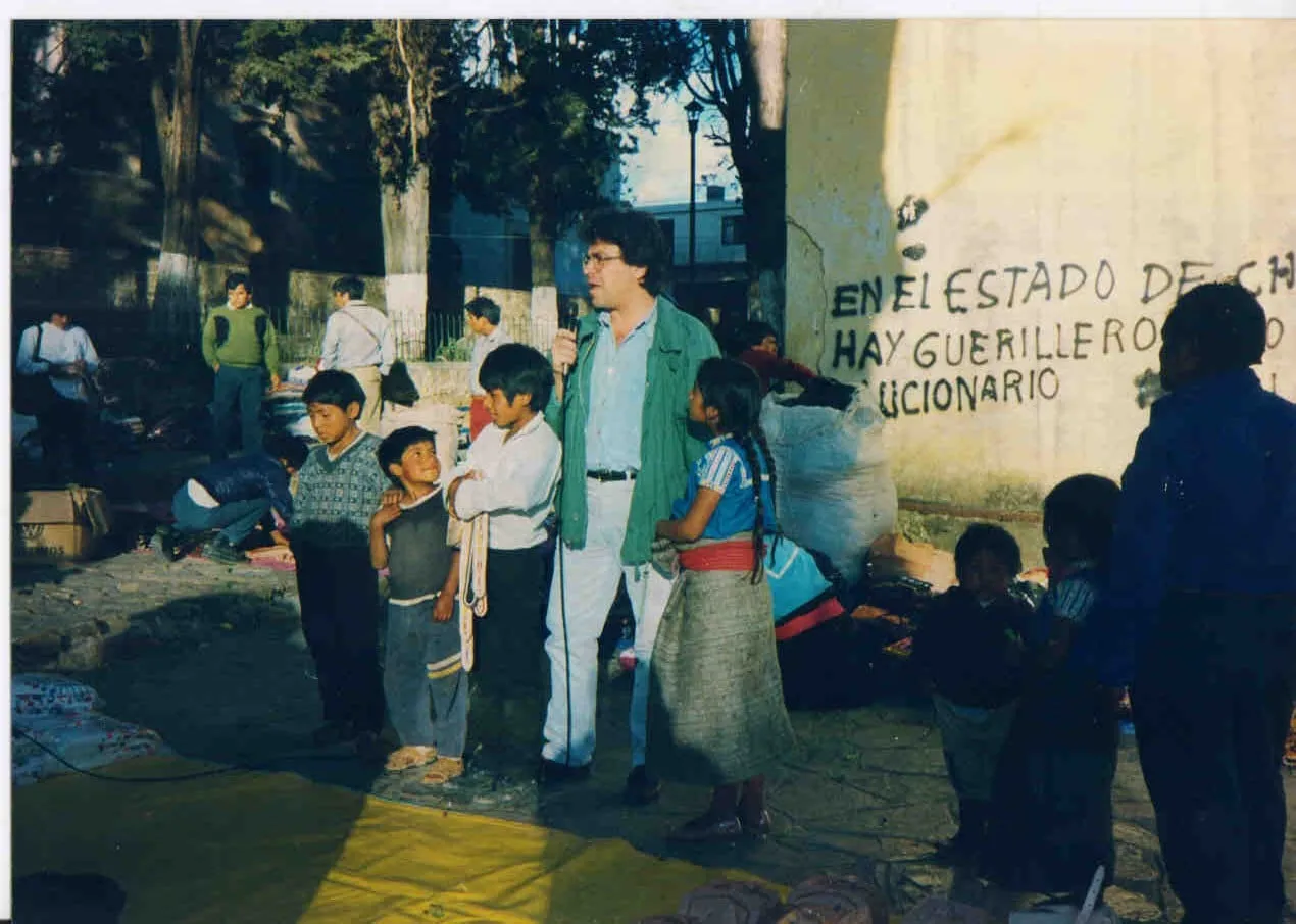 Argyris Dinopoulos standing on paving, in a green jacket, blue shirt, black belt and pale trousers, holding a microphone at face height with its cable falling to the ground. At the left of the frame three boys — one in a knitted pullover, one in a dark sleeveless top, and one in a white shirt holding a woven band. At the right of the frame a girl in a blue embroidered blouse, a red sash, a long grey skirt and a braid down her back, two smaller children, and at the edge of the frame an adult in dark clothes. Behind, a whitewashed wall in black capitals — «EN EL ESTADO DE CH…», «HAY GUERILLERO…» and «…UCIONARIO» — trees, an iron railing, people, sacks and goods spread on the ground; low in the foreground, a yellow tarpaulin.