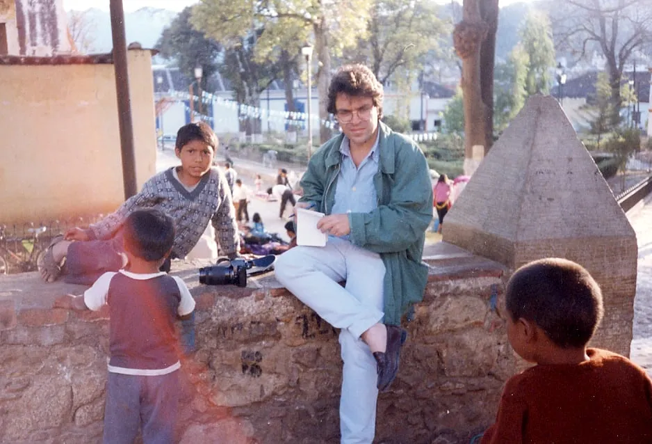 Argyris Dinopoulos sits on a low stone wall with his legs crossed, in a green jacket, blue shirt, pale jeans and dark shoes, holding an open white notepad and a pen, looking at the lens. Beside him, on the wall, a camera with a long lens. At the left a boy in a knitted jumper leans on the wall; further left, lying along the wall, a child in red clothes, bare feet in sandals towards the edge of the frame; in the left foreground, his back to the lens, a small child in a red top with white sleeves, and at the lower right the back of a child's head in a rust-coloured top. Behind, a square with trees, strings of white pennants, a lamp post, low buildings and people sitting with goods on the ground; at the right, a stone pillar with a pyramidal cap.