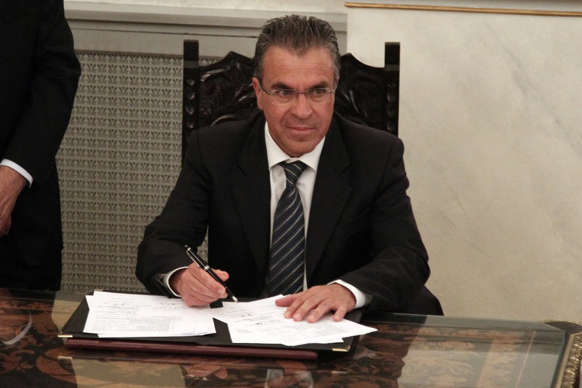 Ο Αργύρης Ντινόπουλος καθισμένος σε γραφείο με σκούρο κοστούμι και γυαλιά, κρατά στυλό πάνω από έγγραφα ακουμπισμένα σε δερμάτινο φάκελο· πίσω του σκαλιστή ξύλινη πλάτη καρέκλας και μαρμάρινος τοίχος, και στο αριστερό άκρο όρθιος άνδρας με σκούρο κοστούμι.