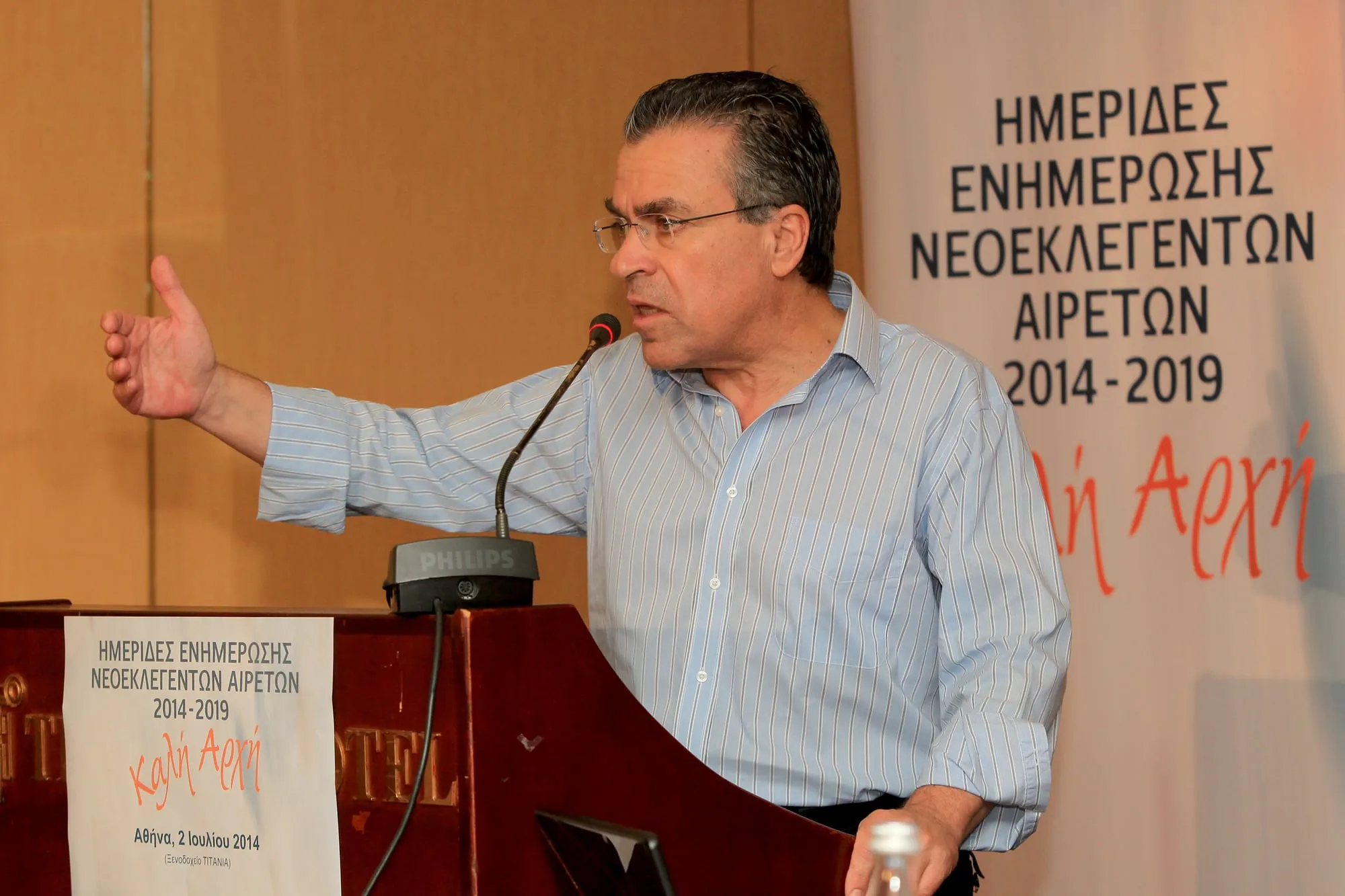 Ο Αργύρης Ντινόπουλος μιλά όρθιος σε αναλόγιο με μικρόφωνο, με γαλάζιο ριγέ πουκάμισο και απλωμένο το δεξί χέρι· πίσω του πανό «ΗΜΕΡΙΔΕΣ ΕΝΗΜΕΡΩΣΗΣ ΝΕΟΕΚΛΕΓΕΝΤΩΝ ΑΙΡΕΤΩΝ 2014-2019 · Καλή Αρχή» και στο αναλόγιο πινακίδα που γράφει «Αθήνα, 2 Ιουλίου 2014».