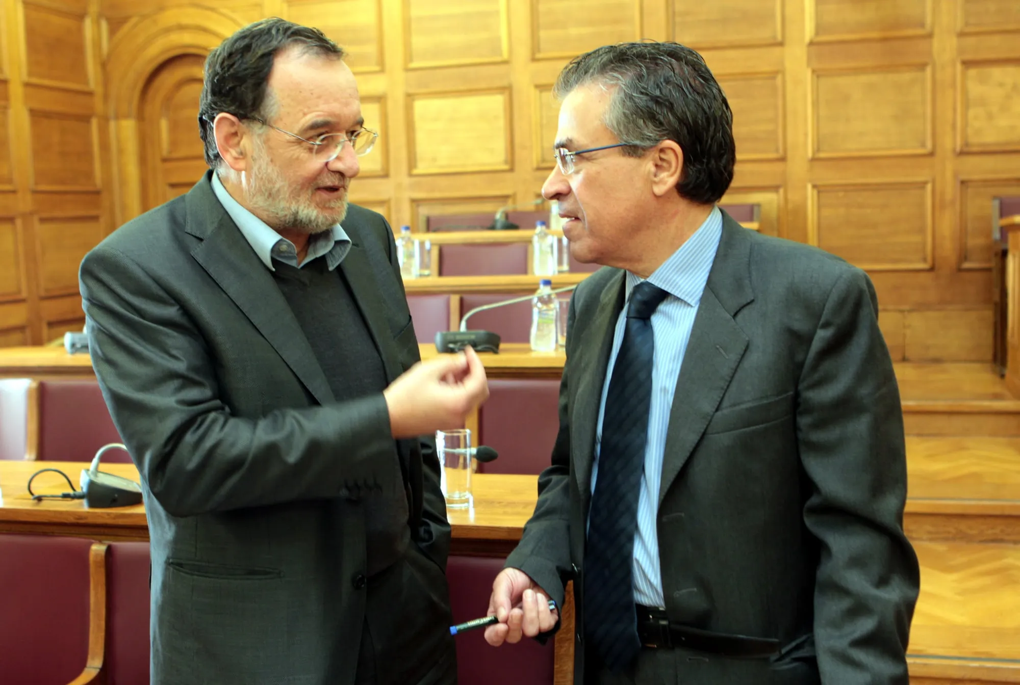 Δύο άνδρες συνομιλούν όρθιοι σε αίθουσα επιτροπής με ξύλινη επένδυση: αριστερά γενειοφόρος άνδρας με γυαλιά και σκούρο σακάκι χωρίς γραβάτα, δεξιά ο Αργύρης Ντινόπουλος με σκούρο κοστούμι και γραβάτα, κρατώντας στυλό· πίσω τους έδρανα με μικρόφωνα.