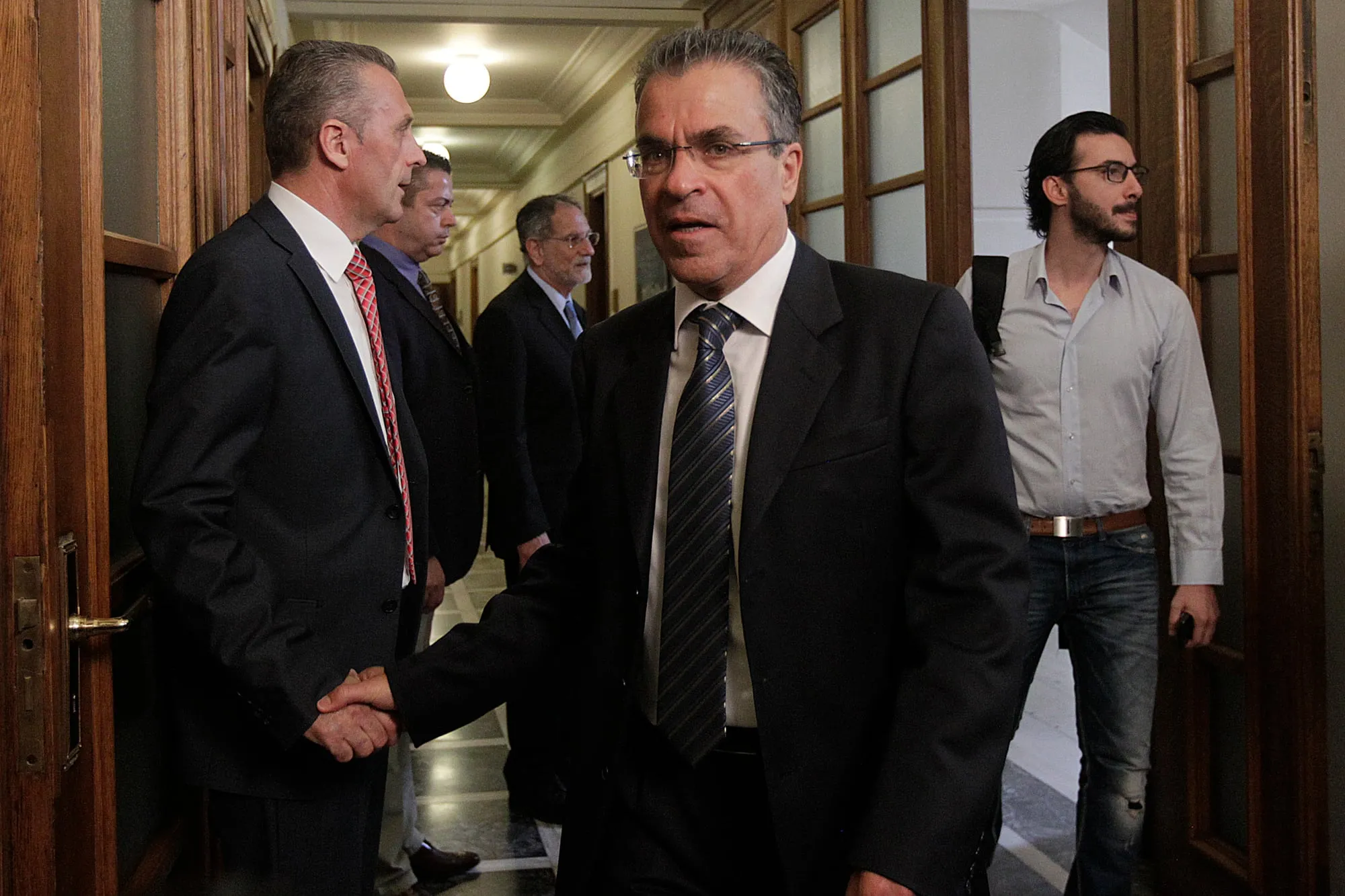 Ο Αργύρης Ντινόπουλος περνά από ανοιχτή ξύλινη πόρτα σε διάδρομο με ξύλινη επένδυση, με σκούρο κοστούμι και ριγέ γραβάτα, δίνοντας το χέρι σε άνδρα στα αριστερά· πίσω τους δύο ακόμη άνδρες με κοστούμια και, δεξιά, νεότερος άνδρας με γκρι πουκάμισο και σακίδιο.