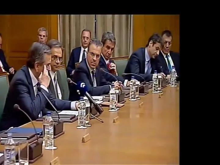 Στιγμιότυπο από τηλεοπτικό πλάνο συνεδρίασης του υπουργικού συμβουλίου: άνδρες με σκούρα κοστούμια καθισμένοι κατά μήκος μακρού γυαλισμένου τραπεζιού με μικρόφωνα, μπουκάλια νερού και ταμπελάκια ονομάτων· μαύρες κάθετες μπάρες στα άκρα του κάδρου.