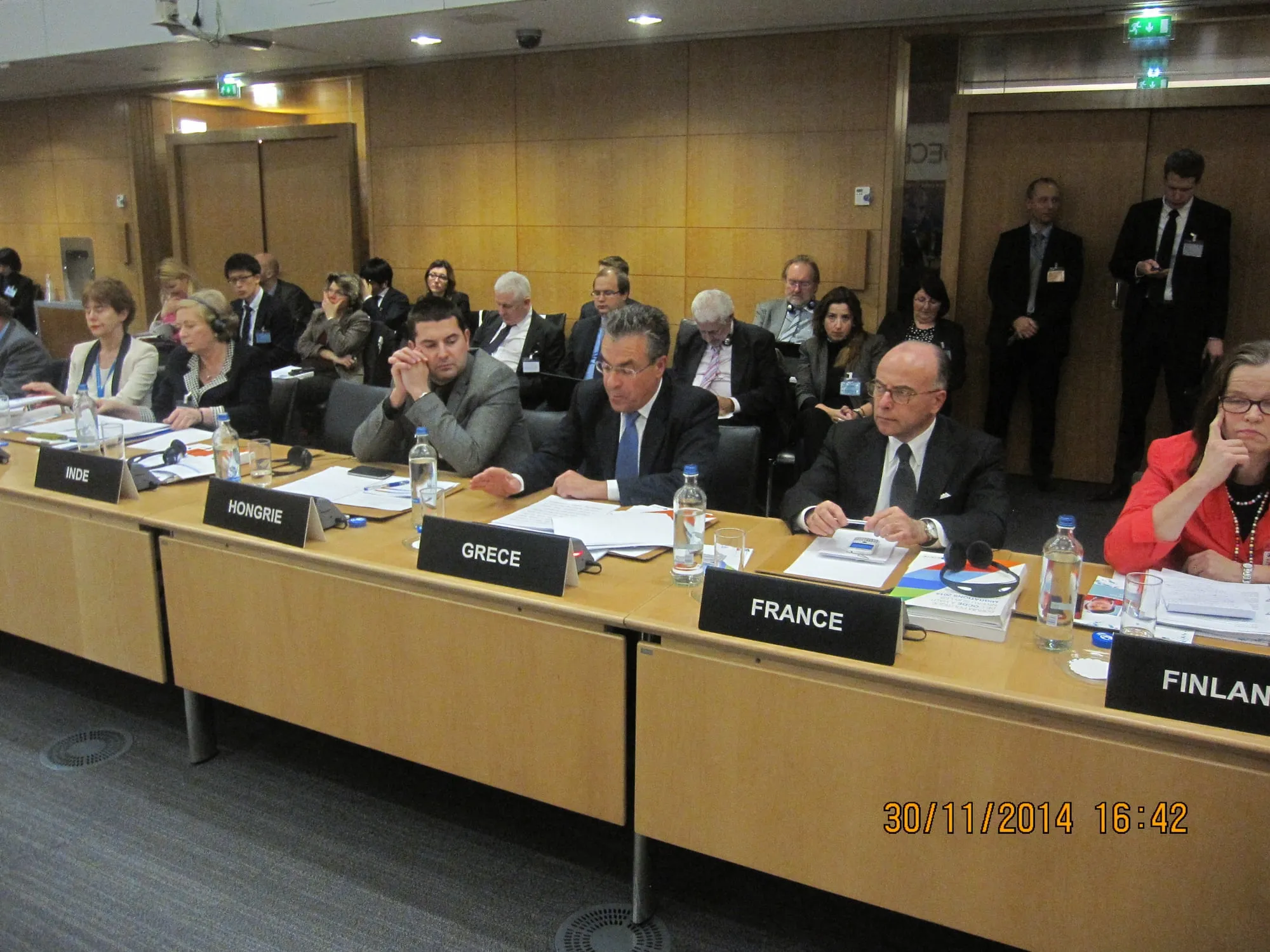 Αίθουσα διεθνούς σύσκεψης: αντιπρόσωποι καθισμένοι πίσω από ξύλινα έδρανα με γαλλόφωνες πινακίδες χωρών — «INDE», «HONGRIE», «GRECE», «FRANCE», «FINLANDE». Ο Αργύρης Ντινόπουλος κάθεται πίσω από την πινακίδα «GRECE» με έγγραφα μπροστά του. Κάτω δεξιά αποτυπωμένη ένδειξη «30/11/2014 16:42».