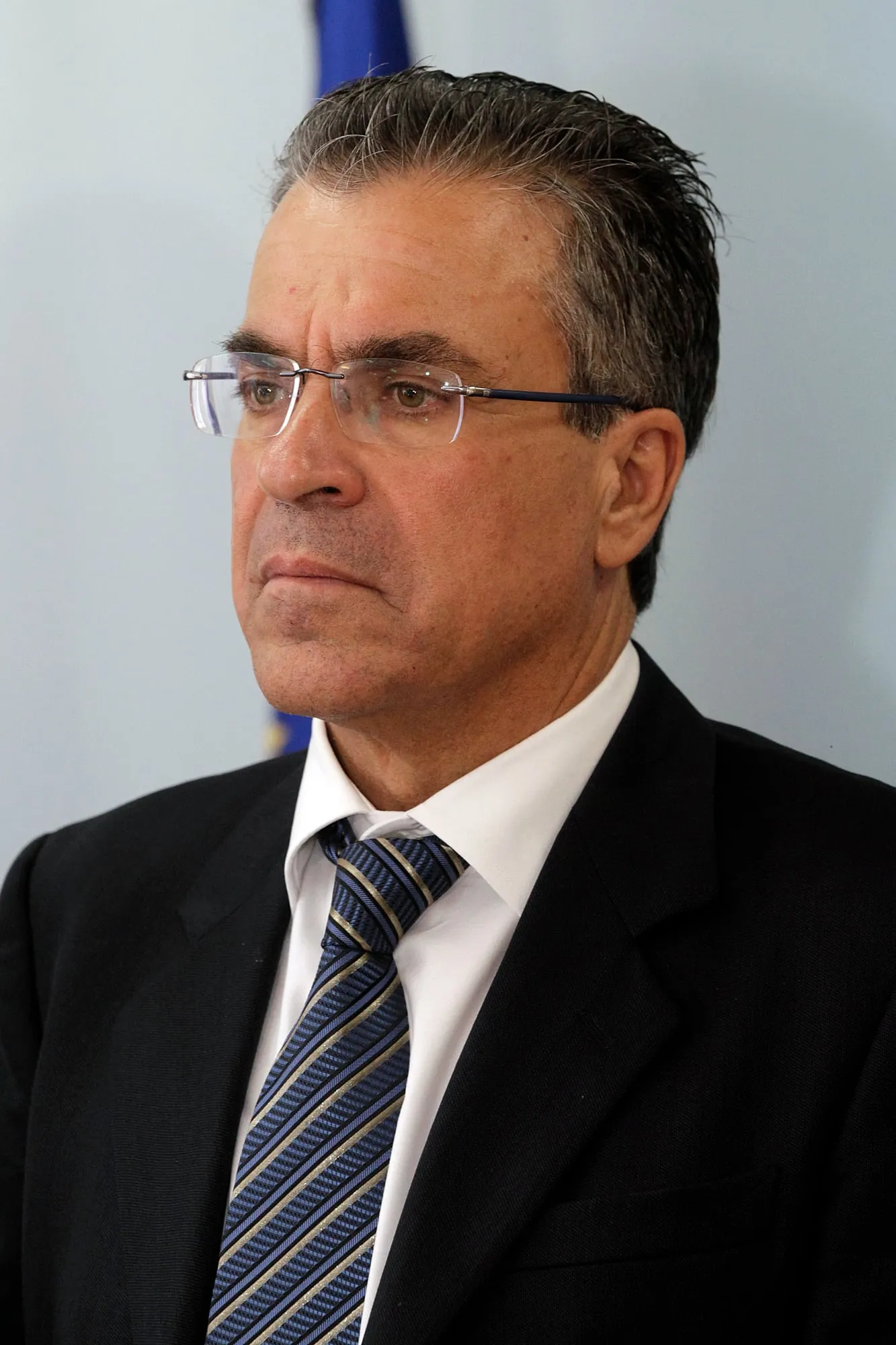 Κοντινό πορτρέτο του Αργύρη Ντινόπουλου με σκούρο κοστούμι, λευκό πουκάμισο και ριγέ γραβάτα σε μπλε και χρυσό, με γυαλιά χωρίς σκελετό· κοιτάζει σοβαρός εκτός κάδρου και πίσω του διακρίνεται θολή γαλάζια σημαία.