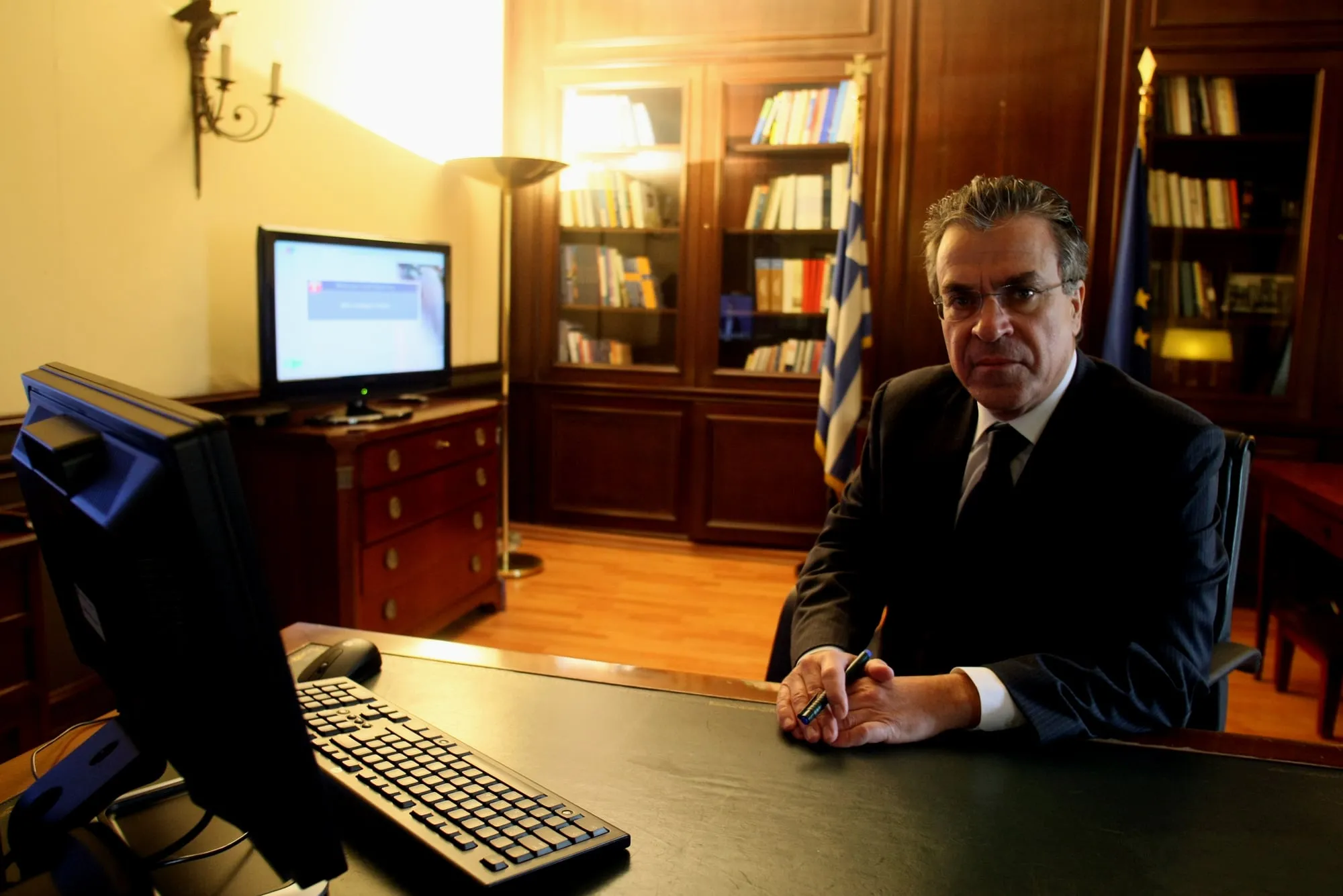 Ο Αργύρης Ντινόπουλος καθισμένος πίσω από μεγάλο γραφείο σε υπουργικό γραφείο με ξύλινη επένδυση, με σκούρο κοστούμι και γυαλιά, κρατώντας στυλό με τα χέρια ενωμένα· μπροστά του οθόνη και πληκτρολόγιο, πίσω του τζαμωτές βιβλιοθήκες, όρθιο φωτιστικό και δύο σημαίες.