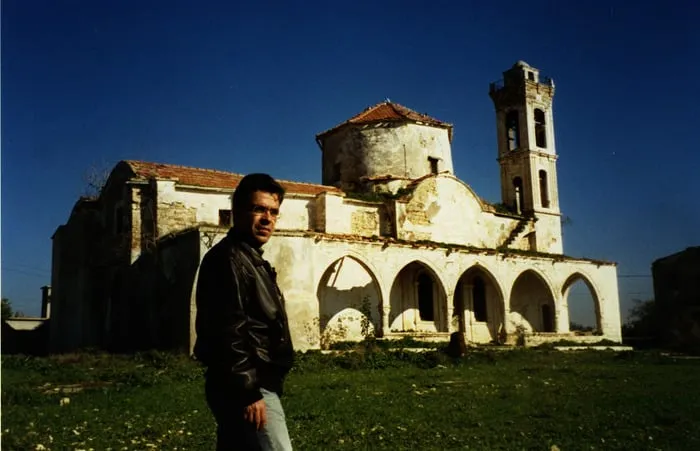Ο ανταποκριτής Αργύρης Ντινόπουλος μπροστά σε ερειπωμένη πέτρινη εκκλησία με ψηλό καμπαναριό κάτω από γαλάζιο ουρανό, κατεχόμενη Κύπρος.