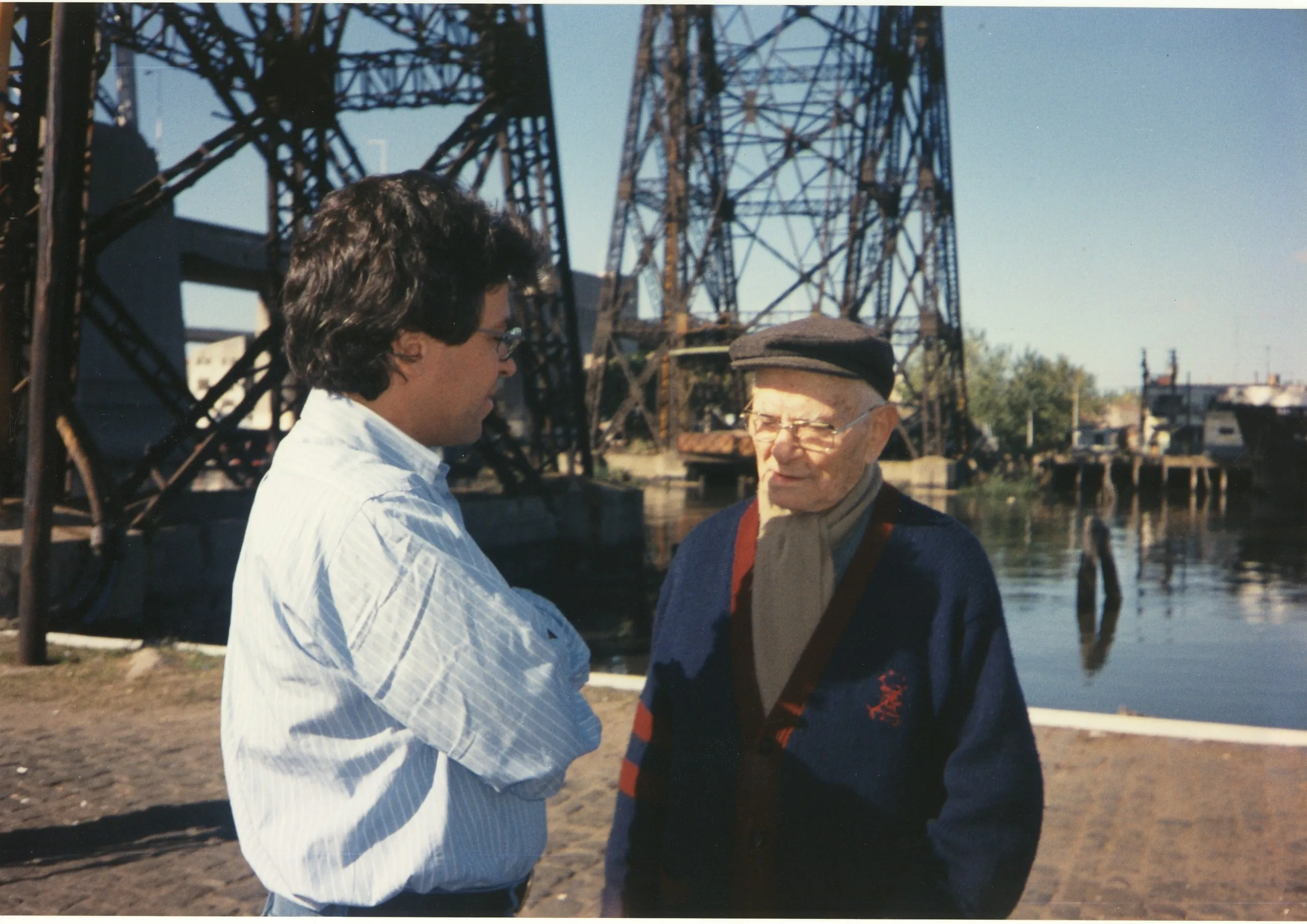 Argyris Dinopoulos, in a striped blue shirt and glasses, stands with his arms folded talking to an elderly man in a flat cap, glasses, a scarf and a blue knitted cardigan with red stripes; behind them a large steel bridge with lattice pylons rises over still water, with low buildings and boats on the far bank; the ground is cobbled and the sky is clear.