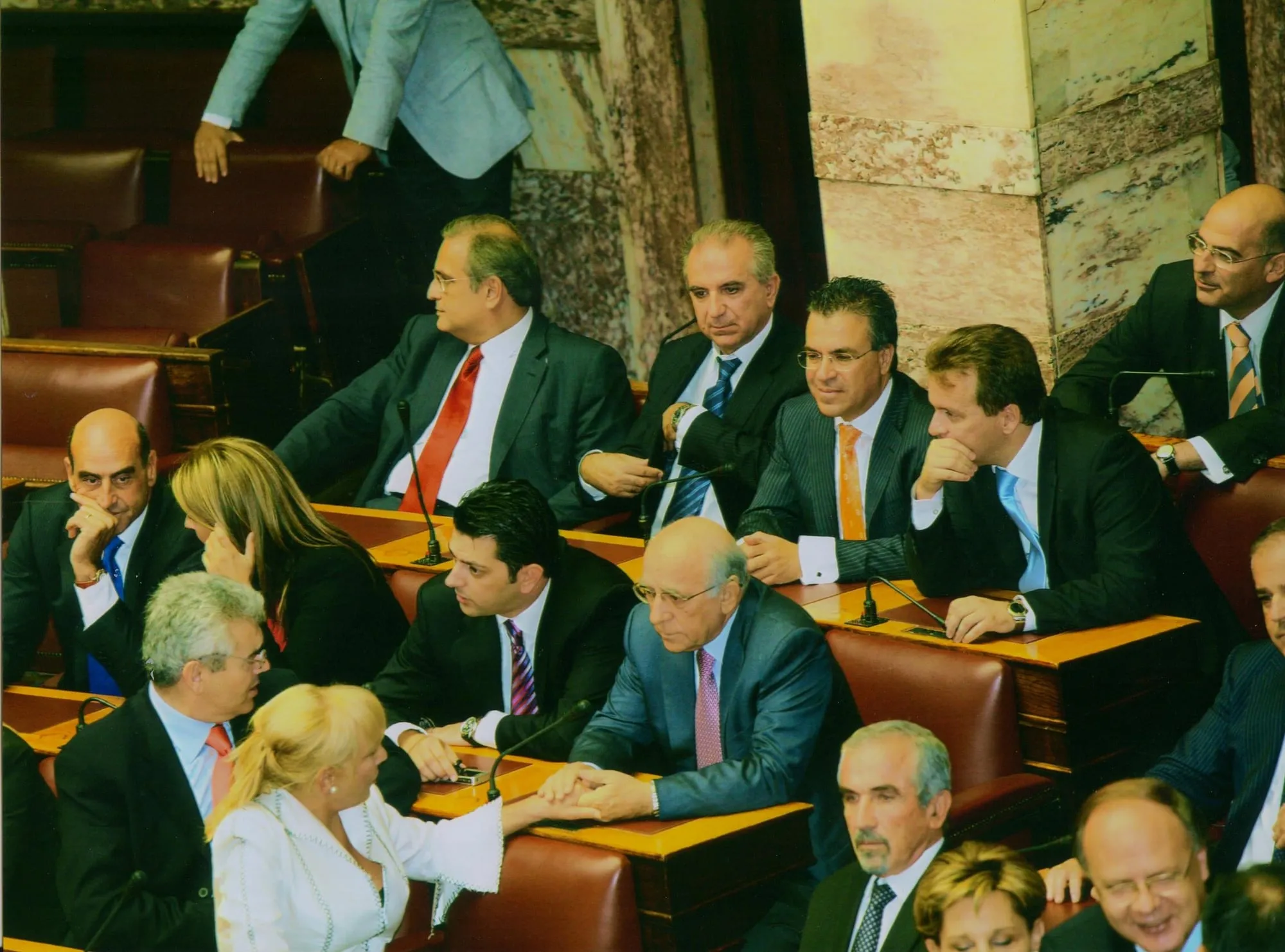 Κλιμακωτά έδρανα με μπορντό δερμάτινες πλάτες και μικρόφωνα στα ξύλινα θρανία, γεμάτα από καθισμένους άνδρες και γυναίκες, κάποιοι από τους οποίους συνομιλούν· στο βάθος μαρμάρινη επένδυση τοίχου.
