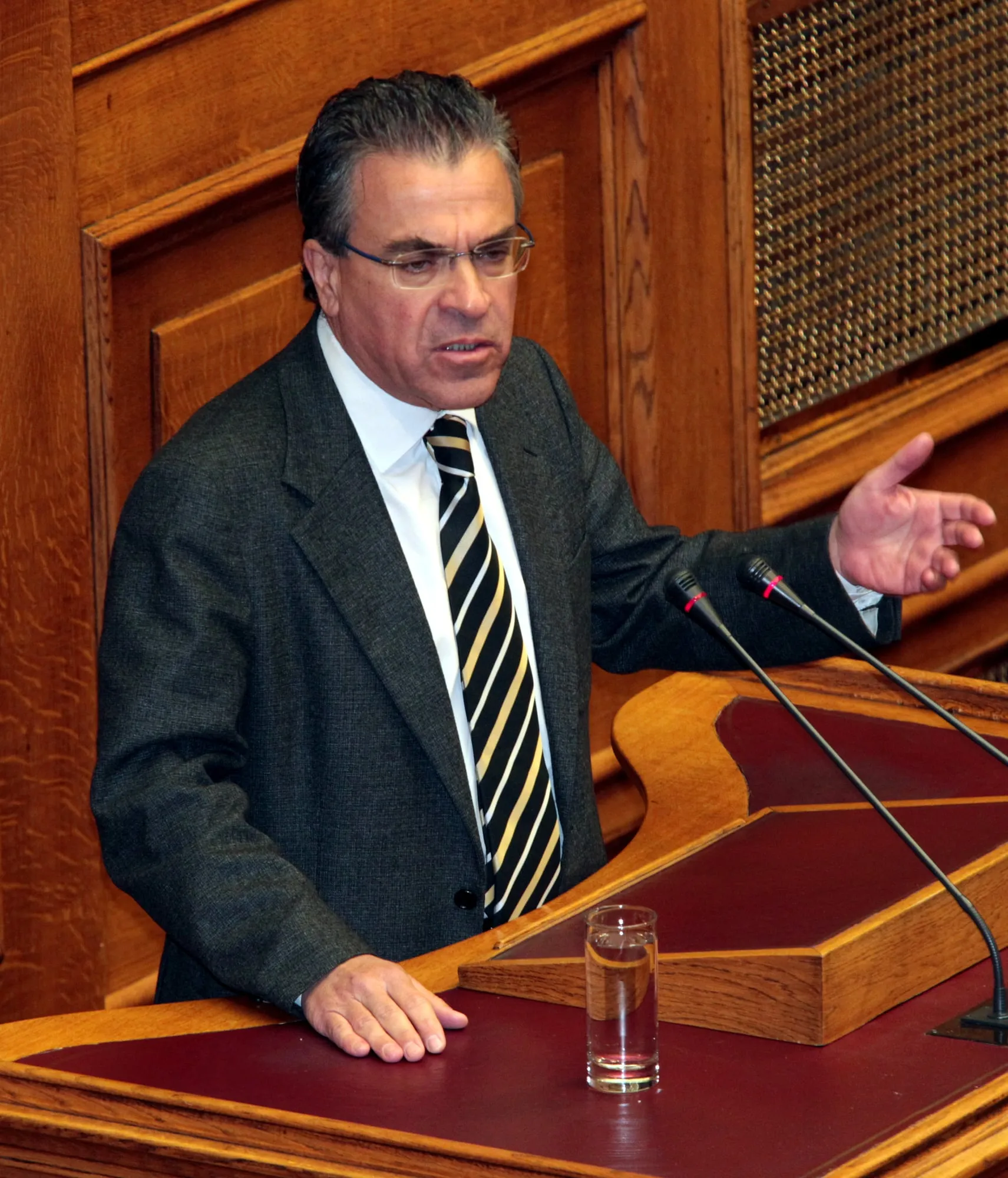 Ο Αργύρης Ντινόπουλος μιλά από ξύλινο βήμα με το ένα χέρι υψωμένο σε χειρονομία, με δύο μικρόφωνα και ένα ποτήρι νερό μπροστά του και ξύλινη επένδυση πίσω του.