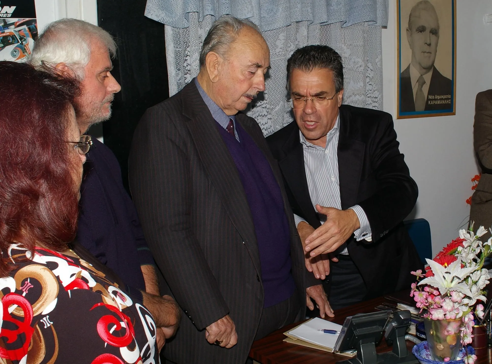 Ο Αργύρης Ντινόπουλος, σκυμμένος μπροστά με ανοιχτή παλάμη, συζητά με ηλικιωμένο άνδρα με ριγέ κοστούμι σε μικρό γραφείο με δαντελένια κουρτίνα· γύρω τους στέκονται κι άλλοι, και στον τοίχο κρέμεται παλιά κορνιζαρισμένη αφίσα «Νέα Δημοκρατία ΚΑΡΑΜΑΝΛΗΣ».