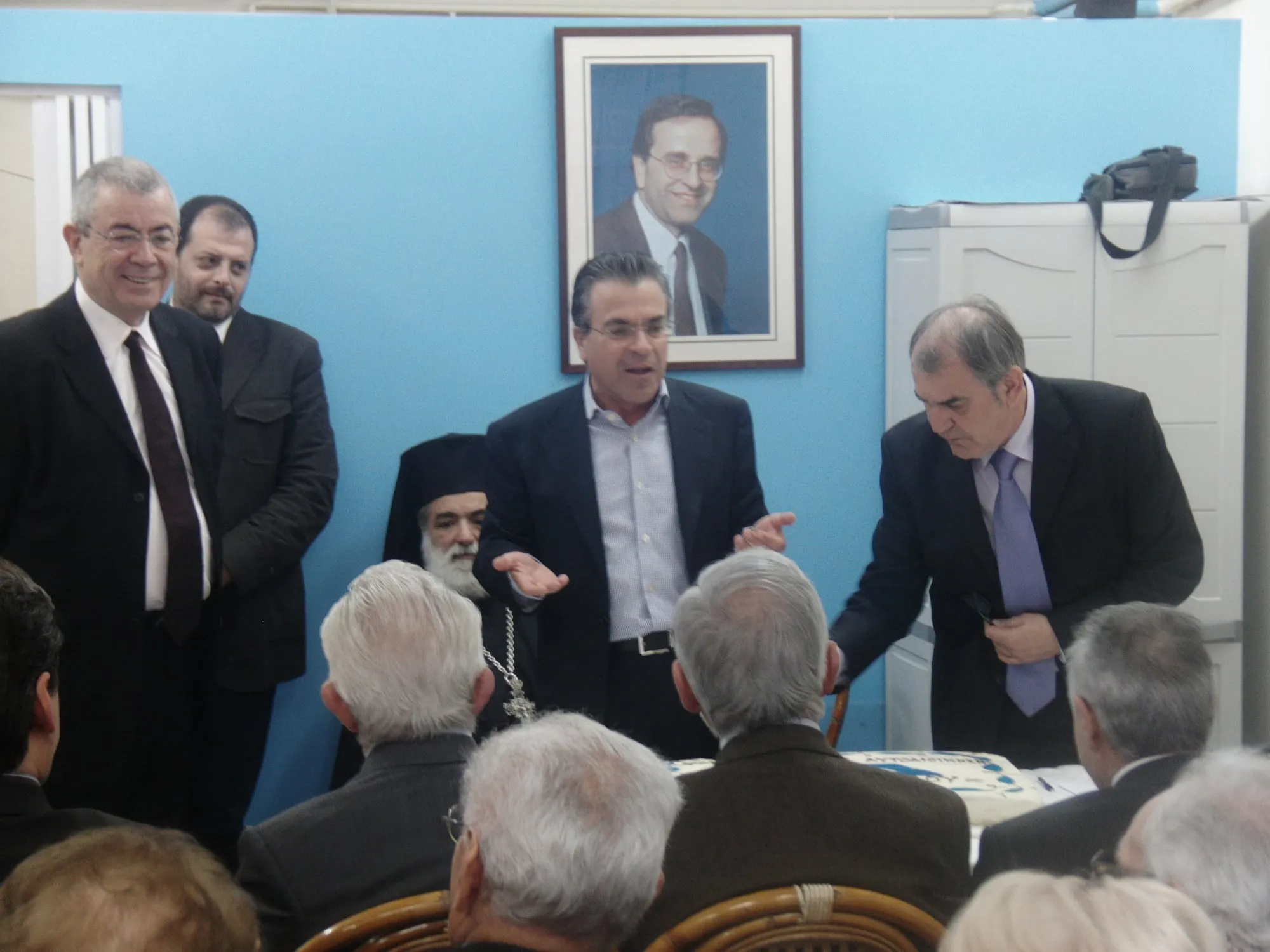 Ο Αργύρης Ντινόπουλος μιλά όρθιος με ανοιχτά χέρια σε αίθουσα με γαλάζιο τοίχο, κάτω από κορνιζαρισμένο πορτρέτο άνδρα με γυαλιά· γύρω του άνδρες με κοστούμια και ένας ορθόδοξος ιερέας, μπροστά καθισμένο ακροατήριο και δεξιά, σε τραπέζι, μεγάλη τούρτα με γαλάζια διακόσμηση.
