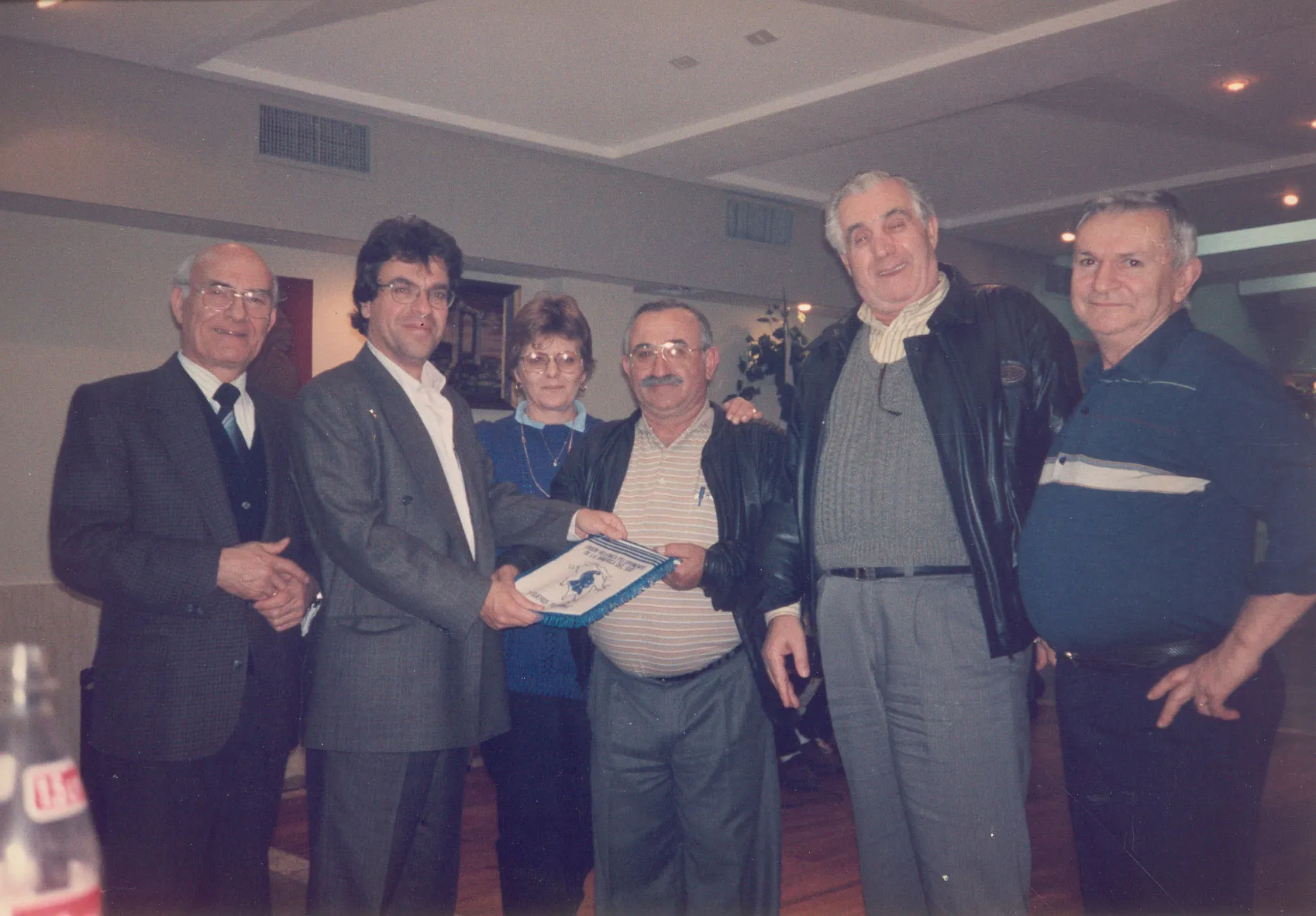 Seven people posing indoors in a room with a wooden floor and a low suspended ceiling; Argyris Dinopoulos, second from the left in a grey jacket, white shirt and glasses, holds a triangular cloth pennant with pale-blue lettering and a fringe together with a moustachioed man; around them stand four more men and a woman in a blue knit, all turned to the lens; at the lower left a bottle stands on a table.