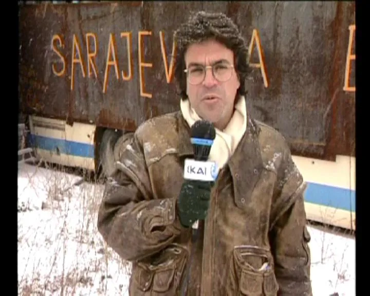 Ο Αργύρης Ντινόπουλος μιλά σε μικρόφωνο με το σήμα του ΣΚΑΪ, μπροστά σε ξεφλουδισμένο τοίχο με τα γράμματα SARAJEVO βαμμένα με μπογιά.